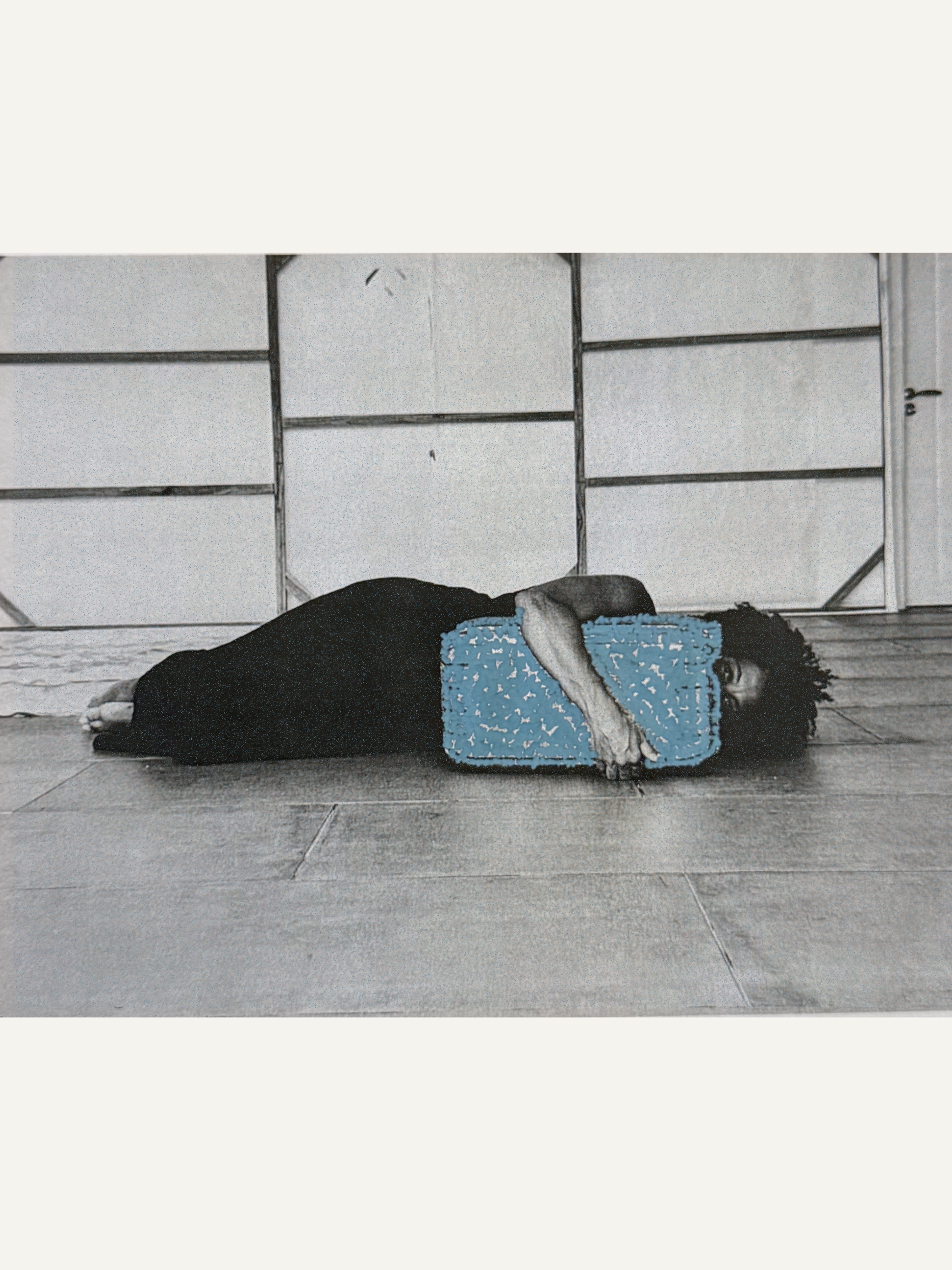 A person lies on a wooden floor, head resting on a bright blue textured rectangular object, in a mostly grayscale image.