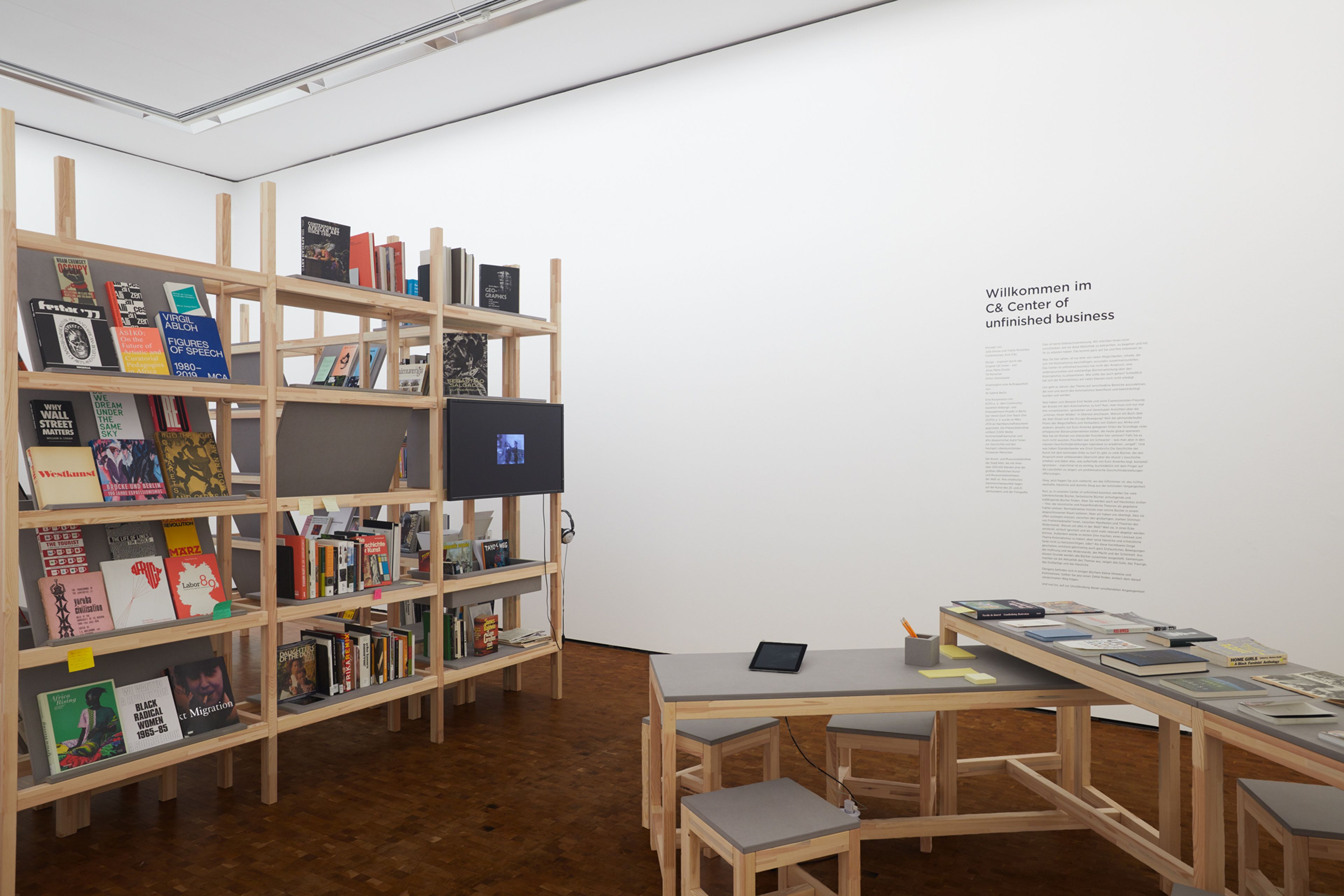 A white room with a tall wooden bookshelf displaying books and a screen, and a long wooden table with stools. Wall text reads, "Willkommen im C& Center of unfinished business."