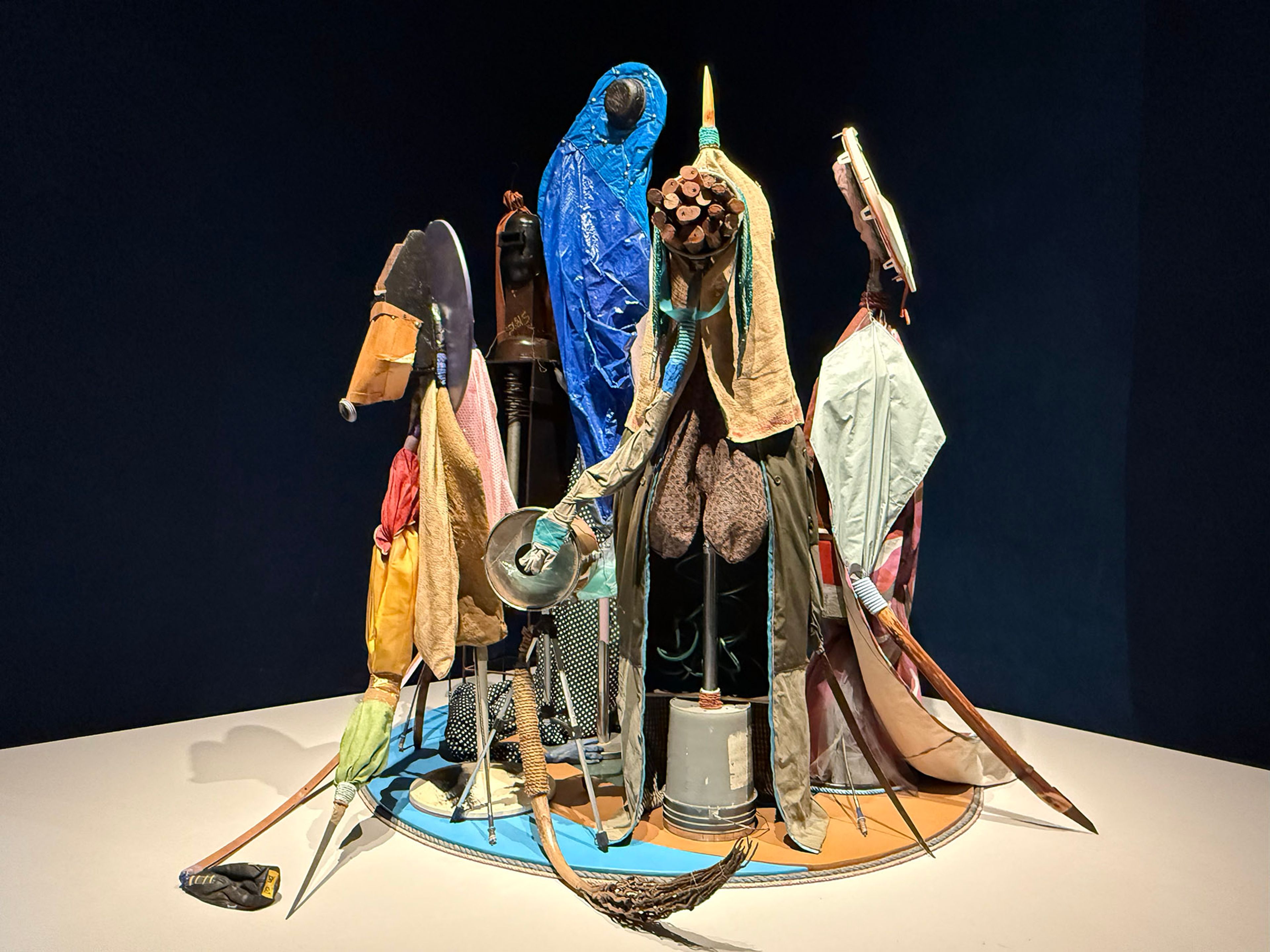 Assemblage sculpture of abstract figures made from colorful fabrics and found objects on a circular base.
