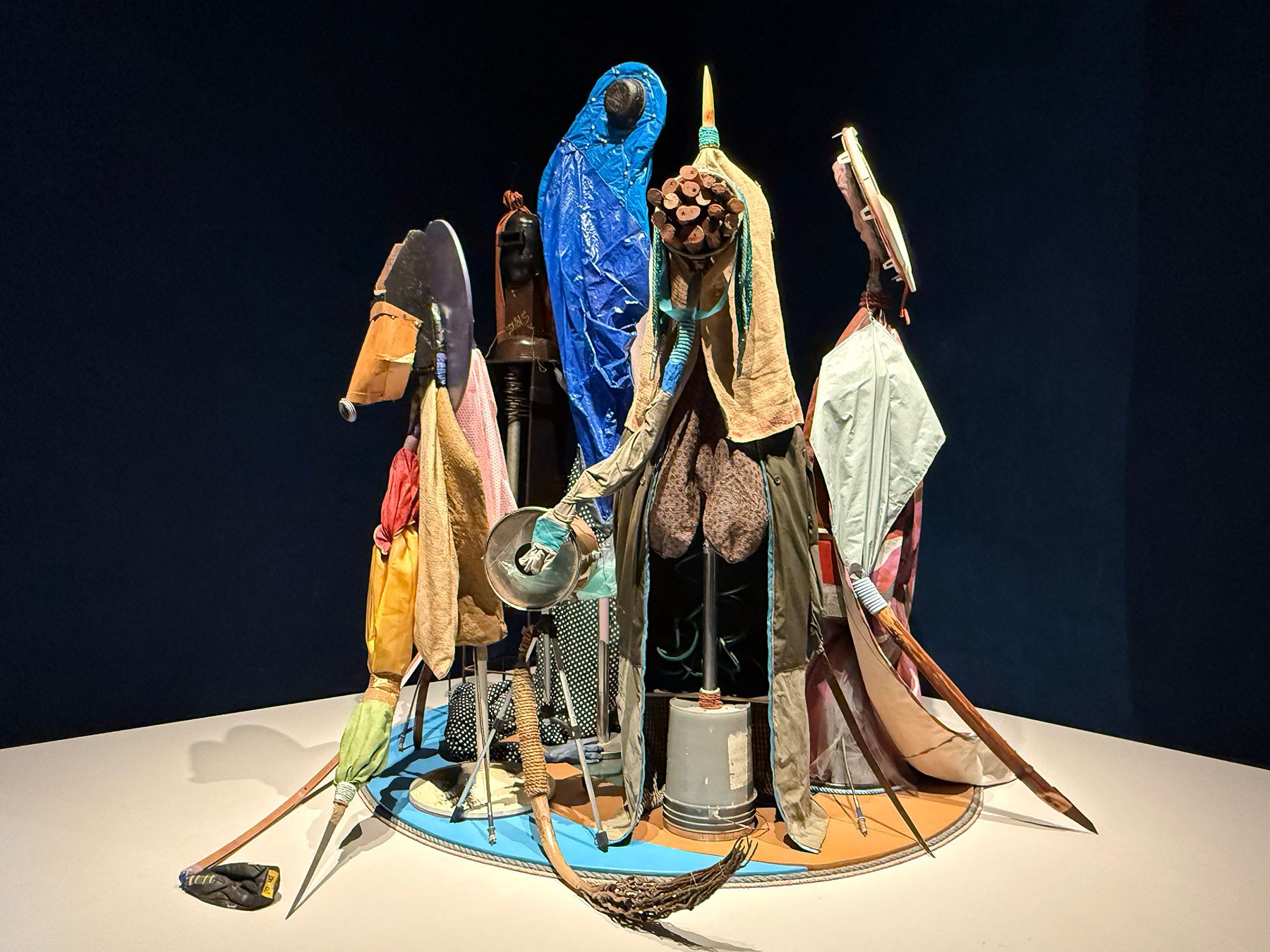 Assemblage sculpture of abstract figures made from colorful fabrics and found objects on a circular base.