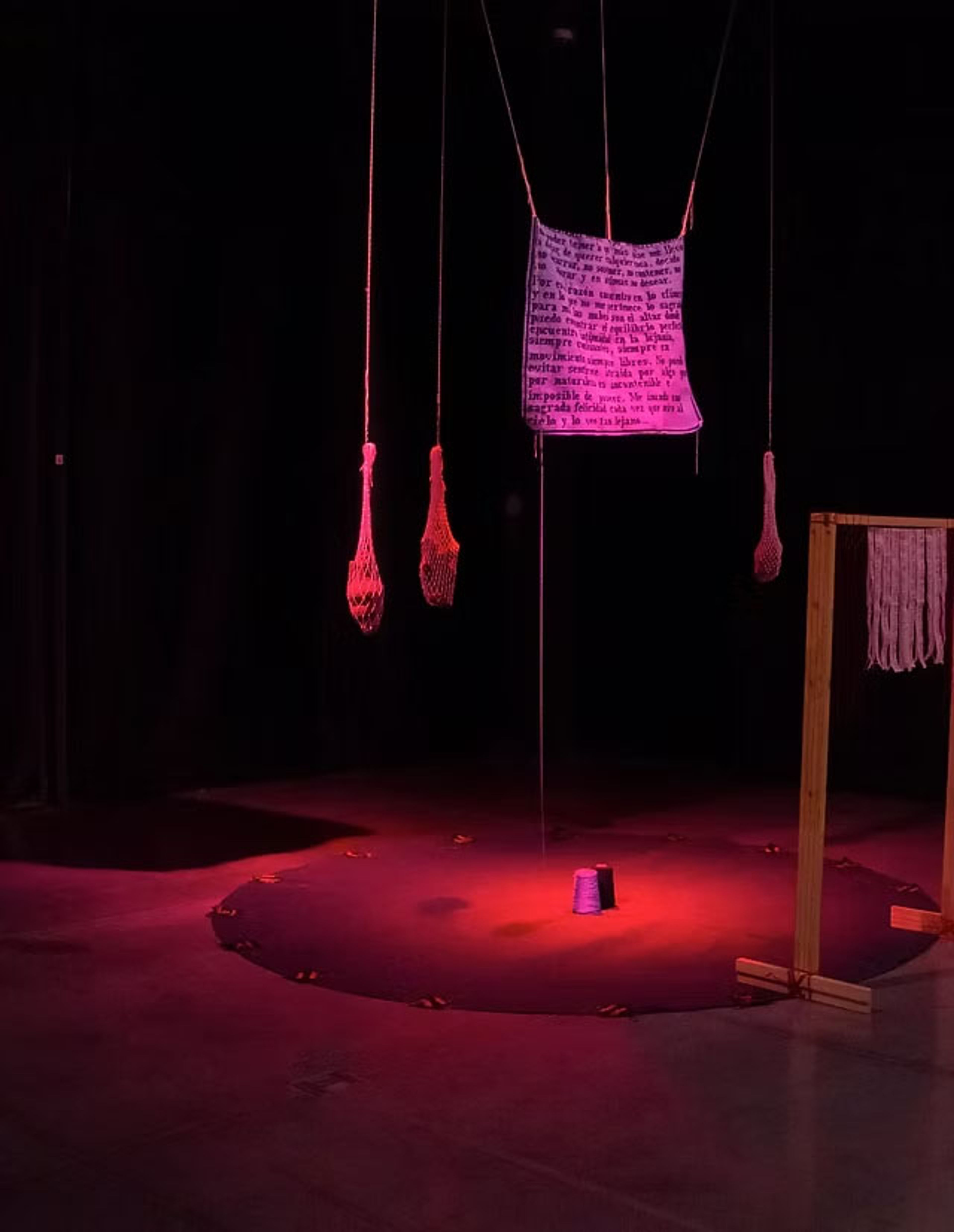 A dimly lit art installation under red and purple light, featuring a hanging text banner, mesh bags, a circle of small shoes around a central purple spool, and a wooden frame with woven pieces.