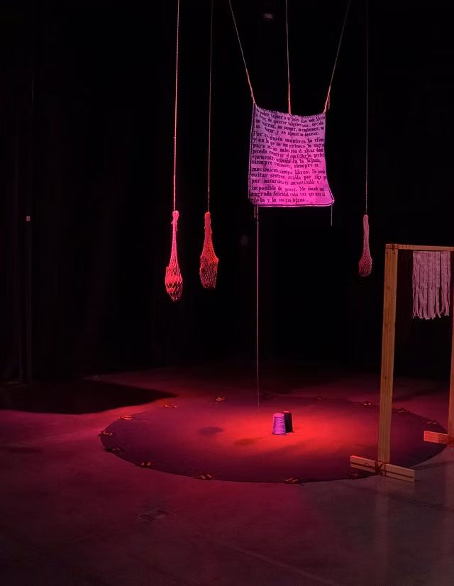 A dimly lit art installation under red and purple light, featuring a hanging text banner, mesh bags, a circle of small shoes around a central purple spool, and a wooden frame with woven pieces.