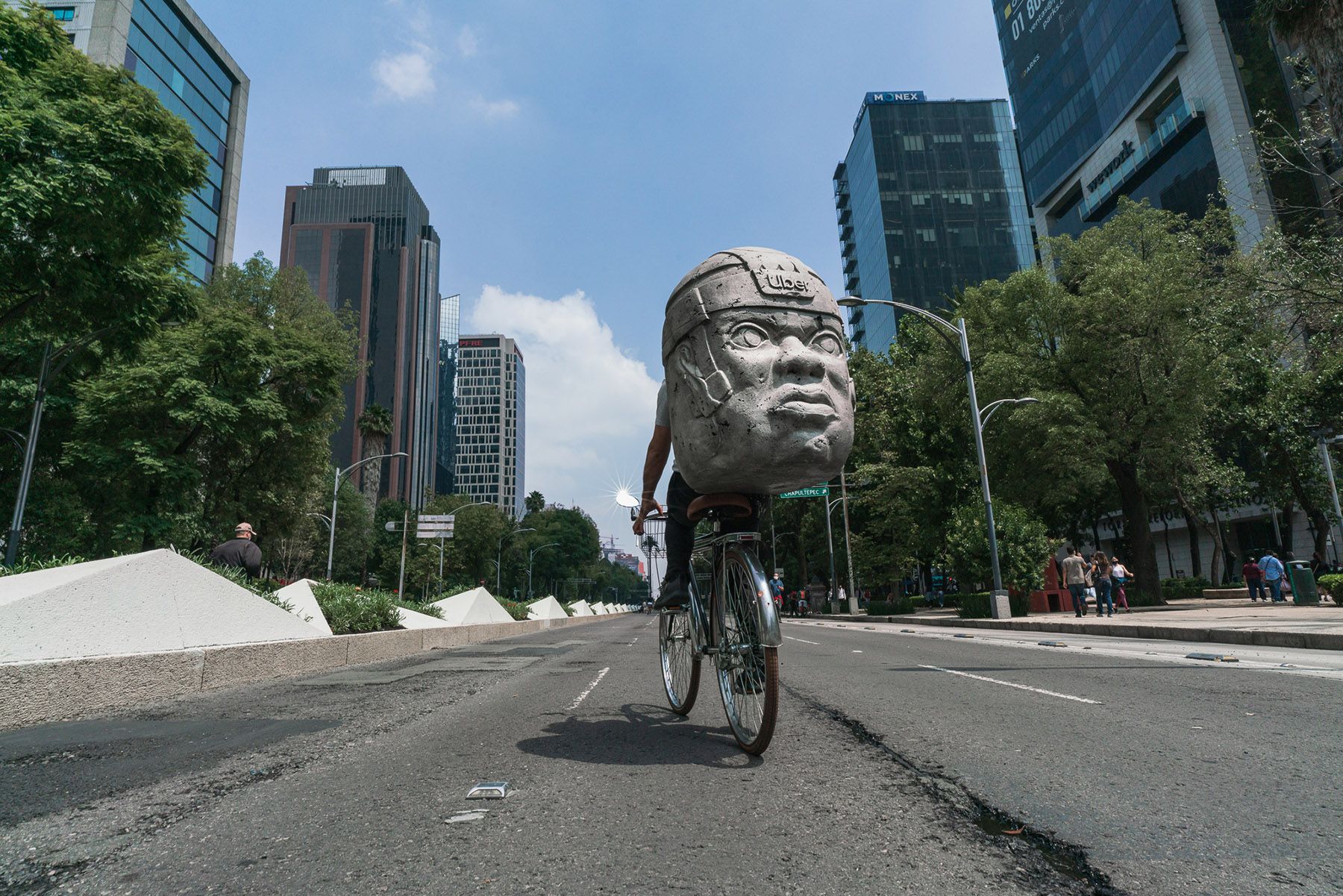 A person cycles down a wide city street, carrying a large replica of an Olmec colossal head on their back.
