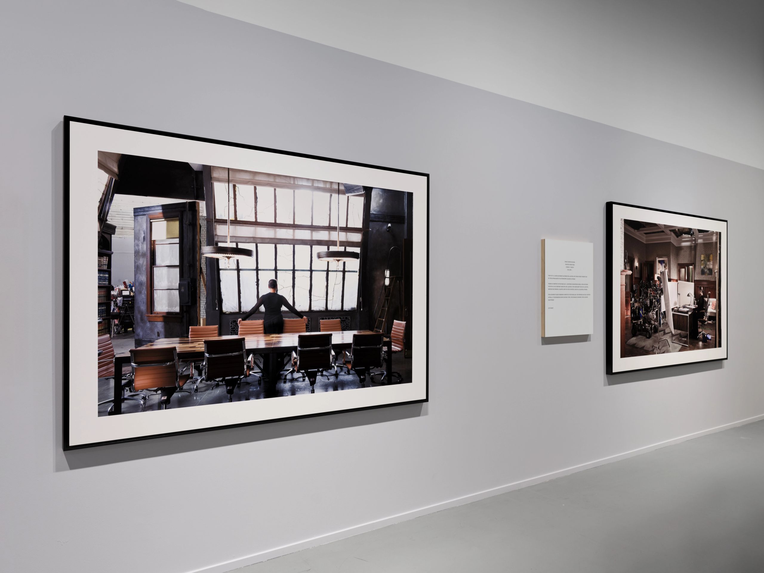 Two framed photographs hang on a grey gallery wall; the larger left photo shows a person standing in a modern conference room with large windows.