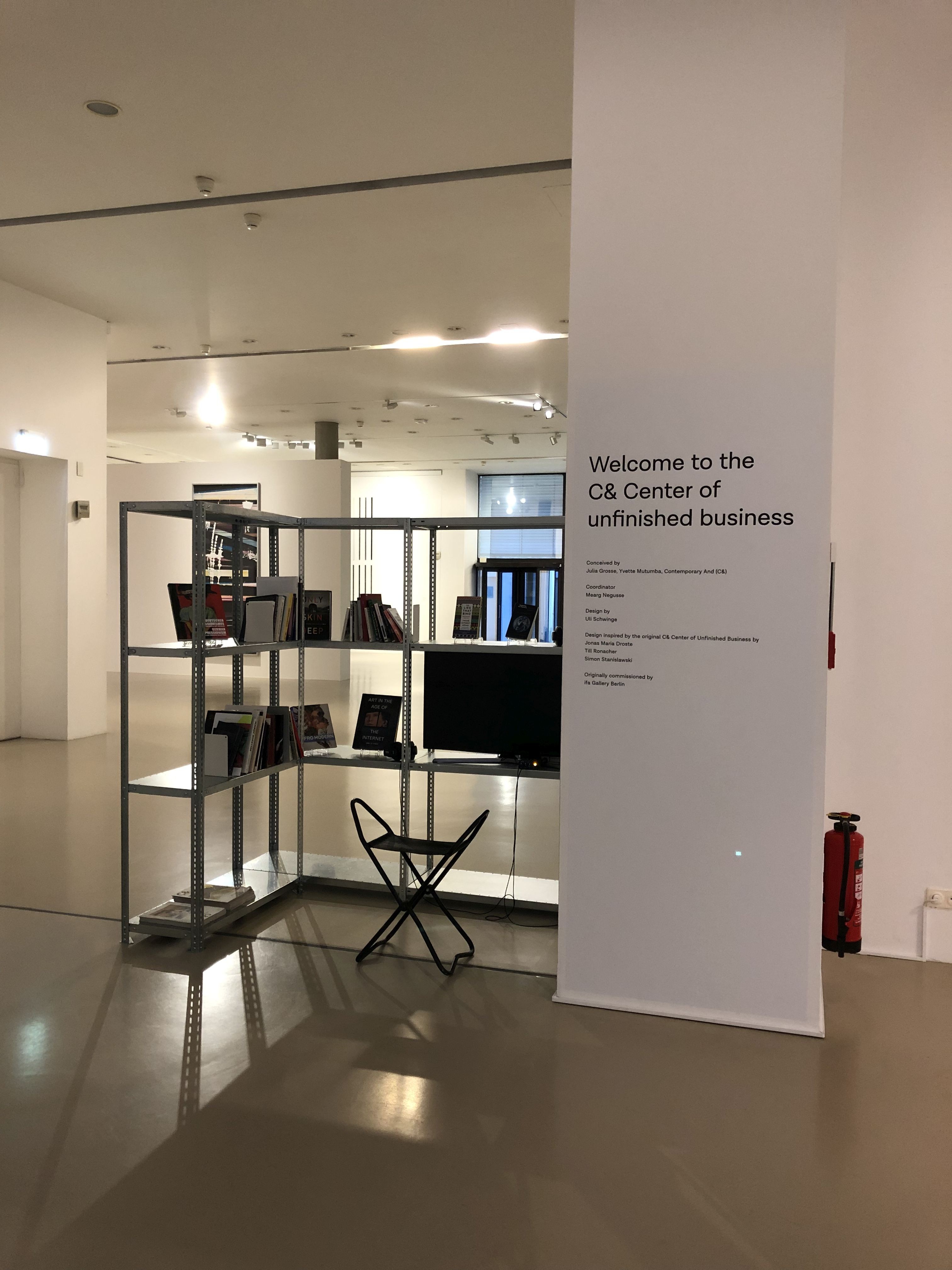 An exhibition space features a white banner reading "Welcome to the C& Center of unfinished business" next to an L-shaped metal shelf with books, frames, and a TV, with a black folding stool in front.