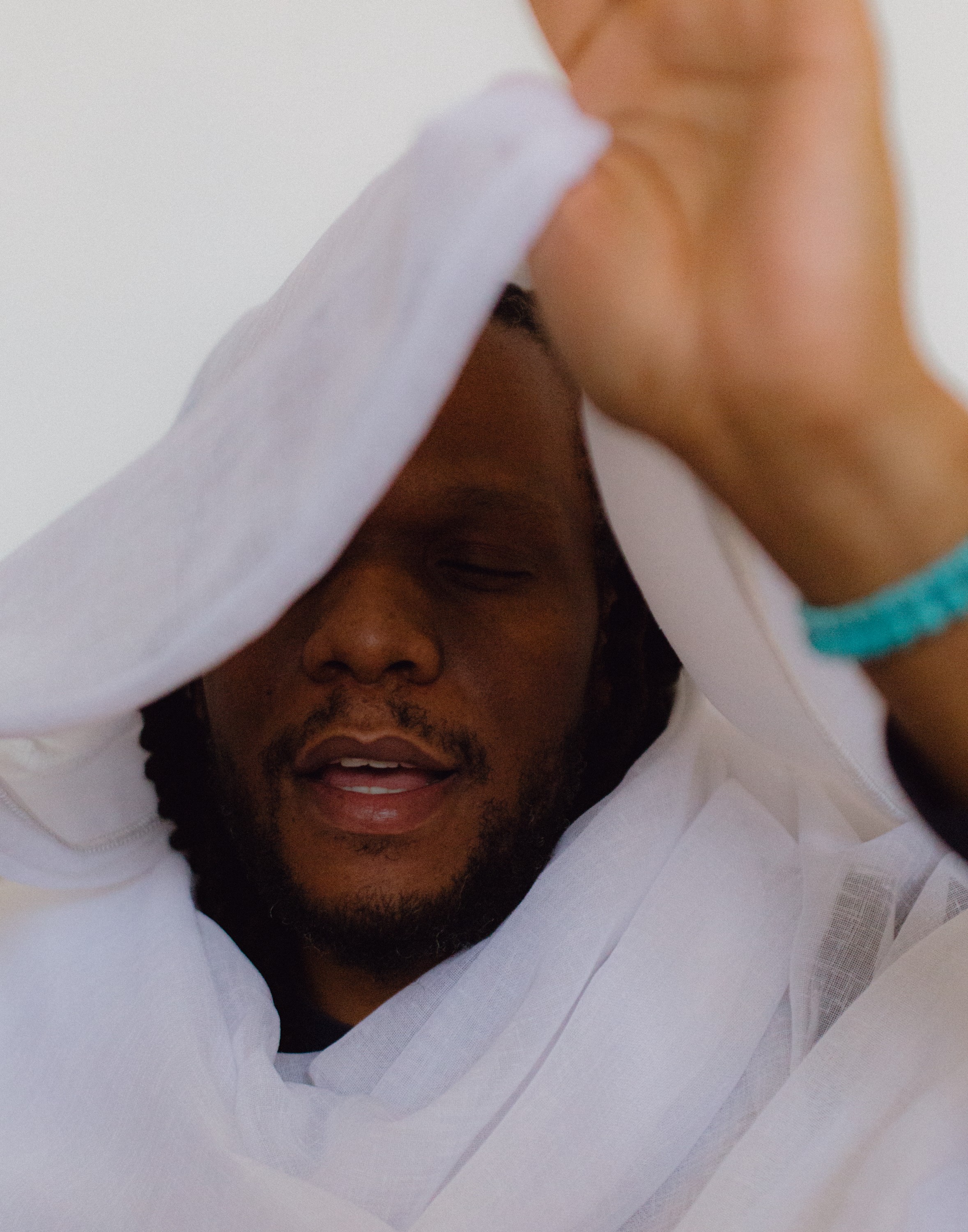 A person with dark skin and dreadlocks, draped in white fabric, eyes closed, with a raised hand wearing a turquoise bracelet.