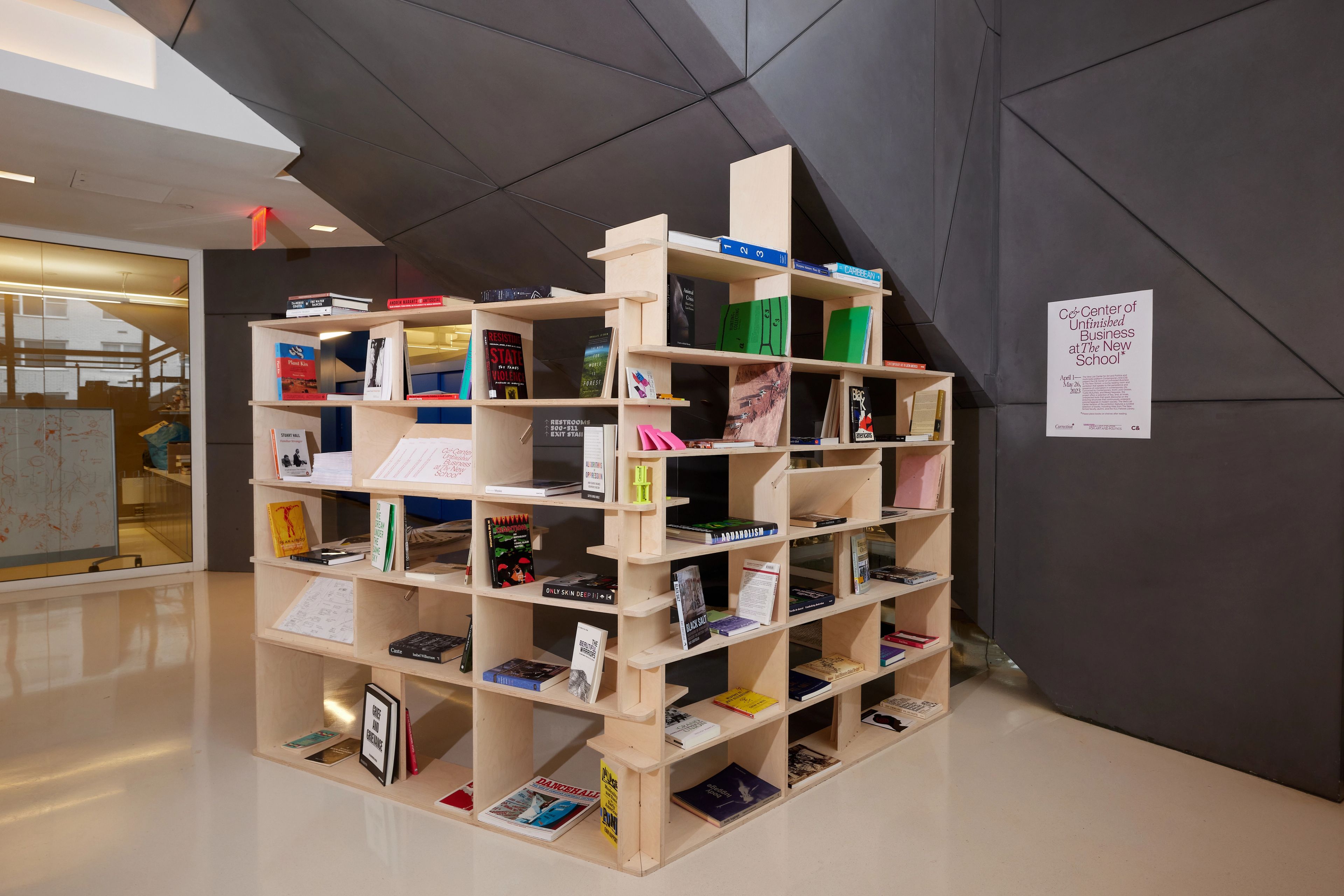 A large, light wood modular bookshelf filled with books stands in a modern room with a dark, geometrically paneled wall.