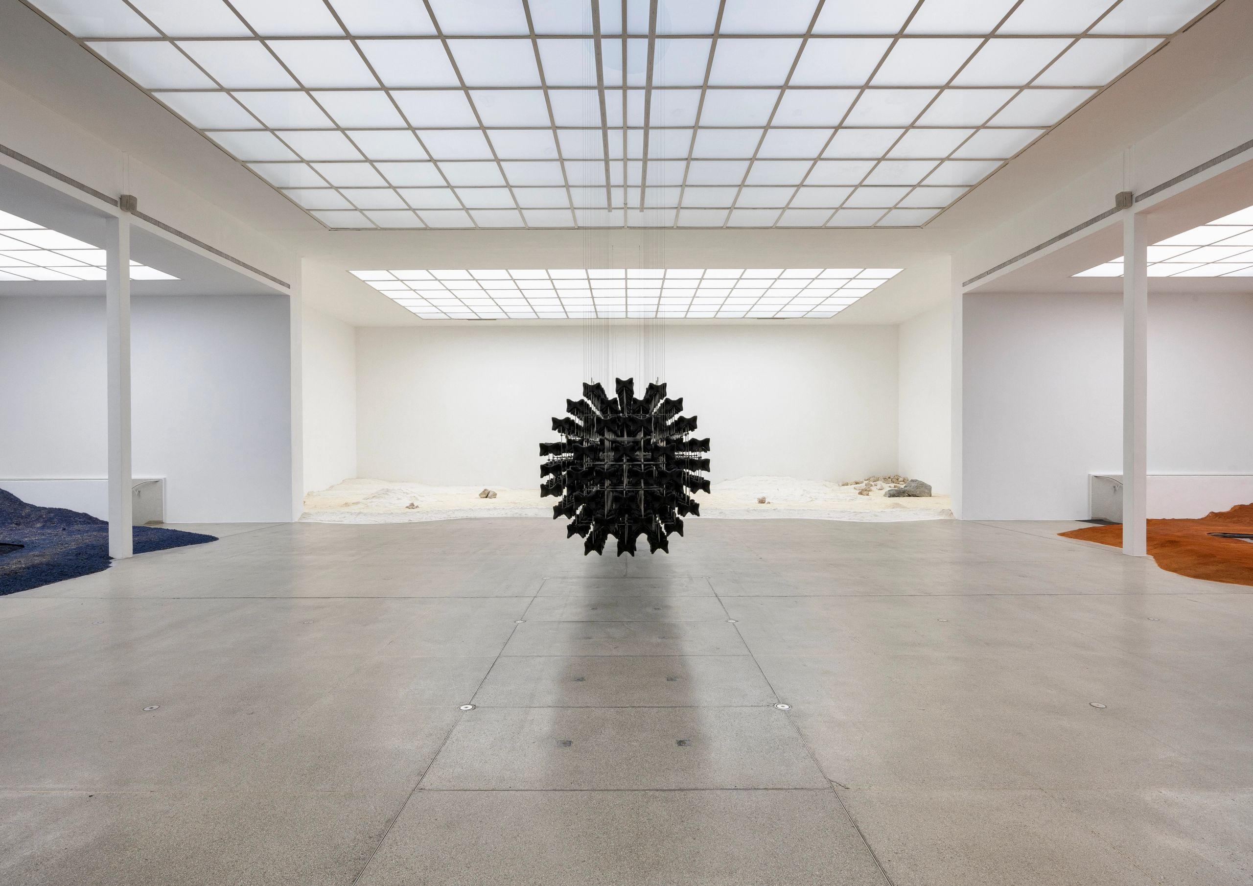 A modern art gallery features a large, spiky black sphere hanging centrally, surrounded by floor installations of wavy blue fabric, brown earth, and light sand.