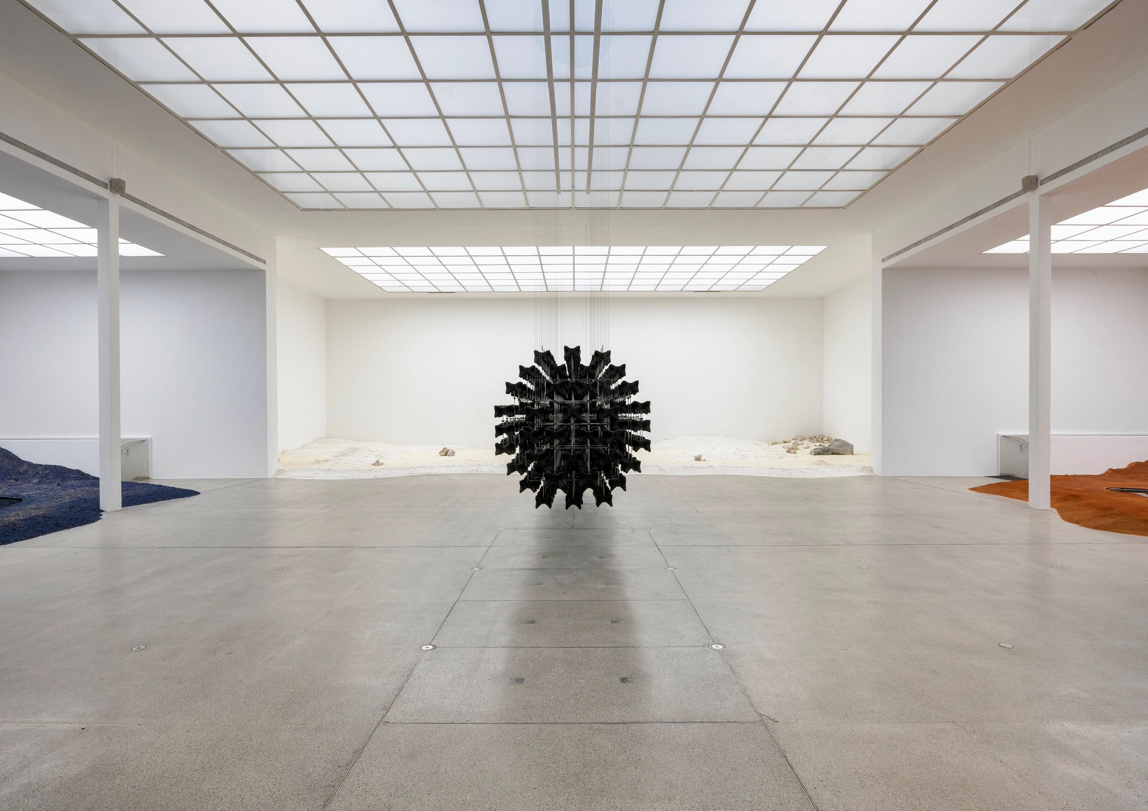 A modern art gallery features a large, spiky black sphere hanging centrally, surrounded by floor installations of wavy blue fabric, brown earth, and light sand.