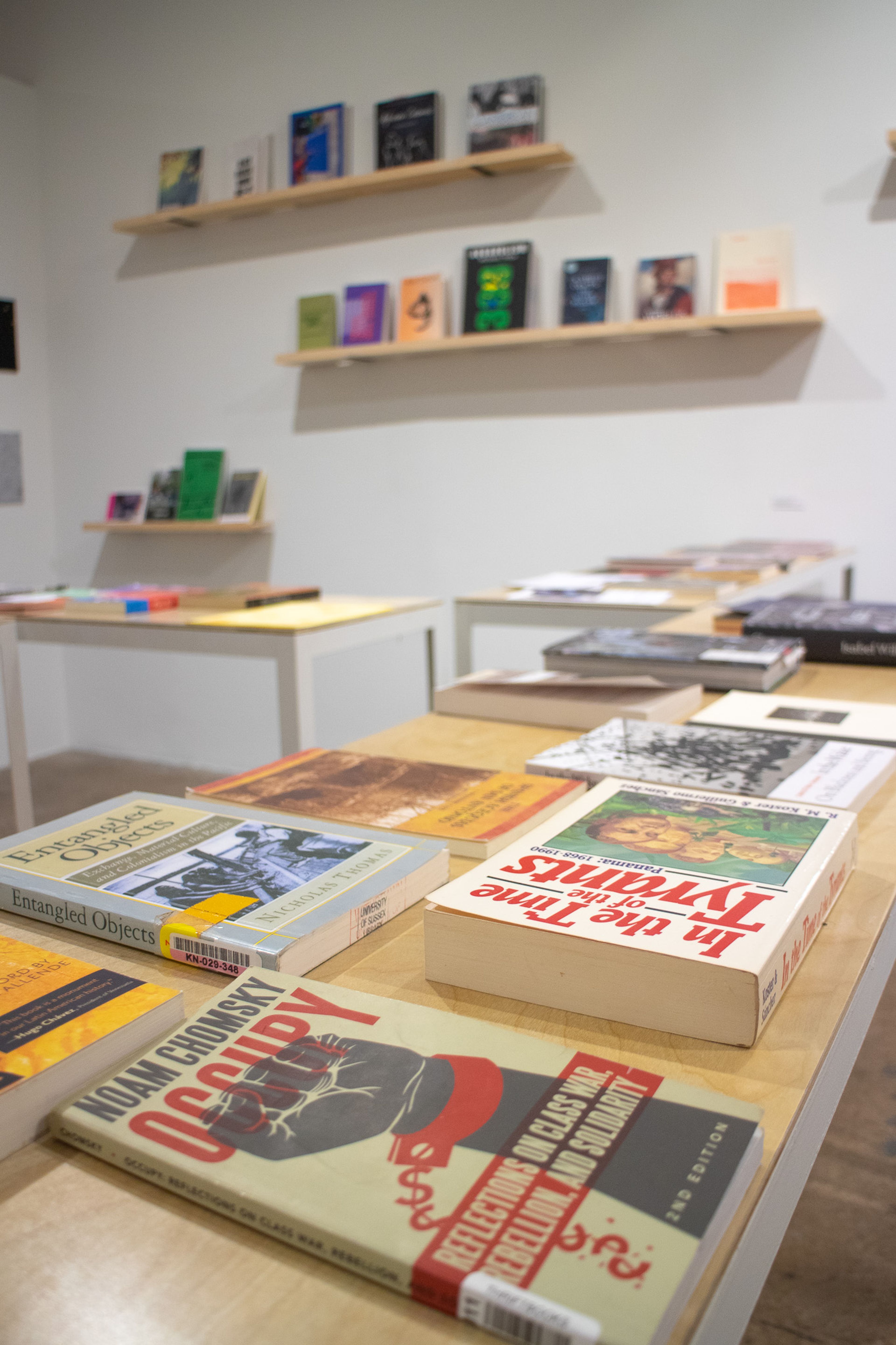 A display of books on light-colored tables in the foreground and on shelves against a white wall in the background. Titles like "Noam Chomsky Occupy" are visible.