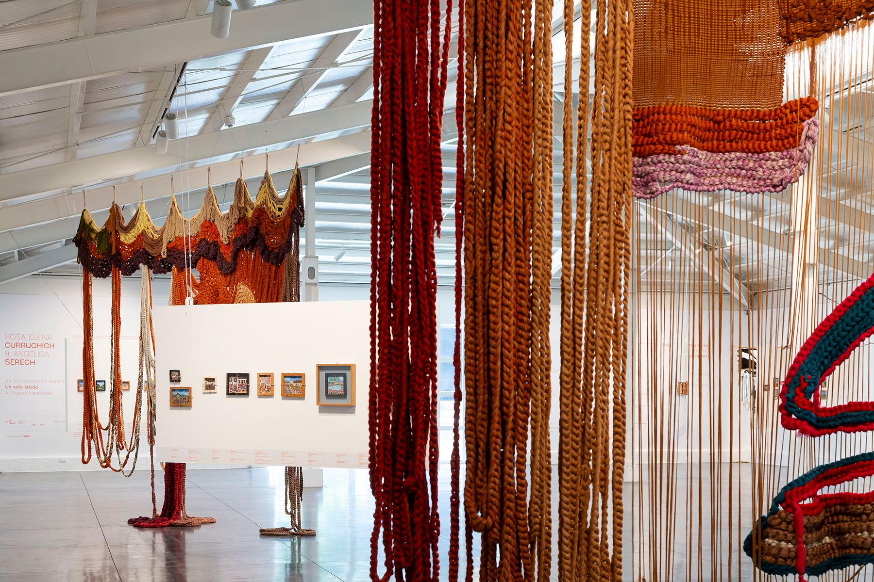 Textile art installation in a gallery, featuring long hanging red and brown ropes and a wavy crocheted piece.