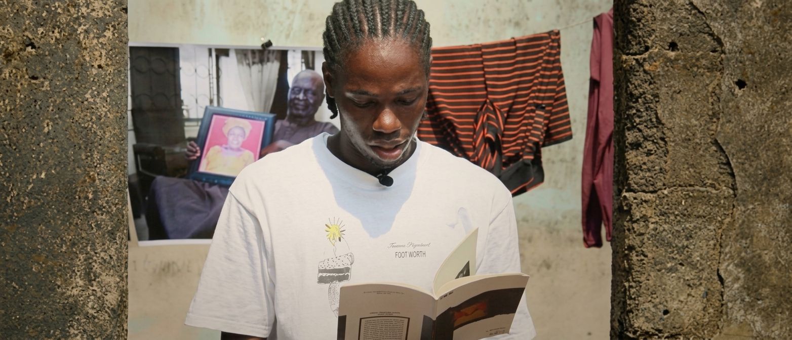 A Black man with braided hair reads a book, wearing a lapel mic. Behind him, a large photo of an older man holding a framed portrait hangs on the wall, next to hanging laundry.