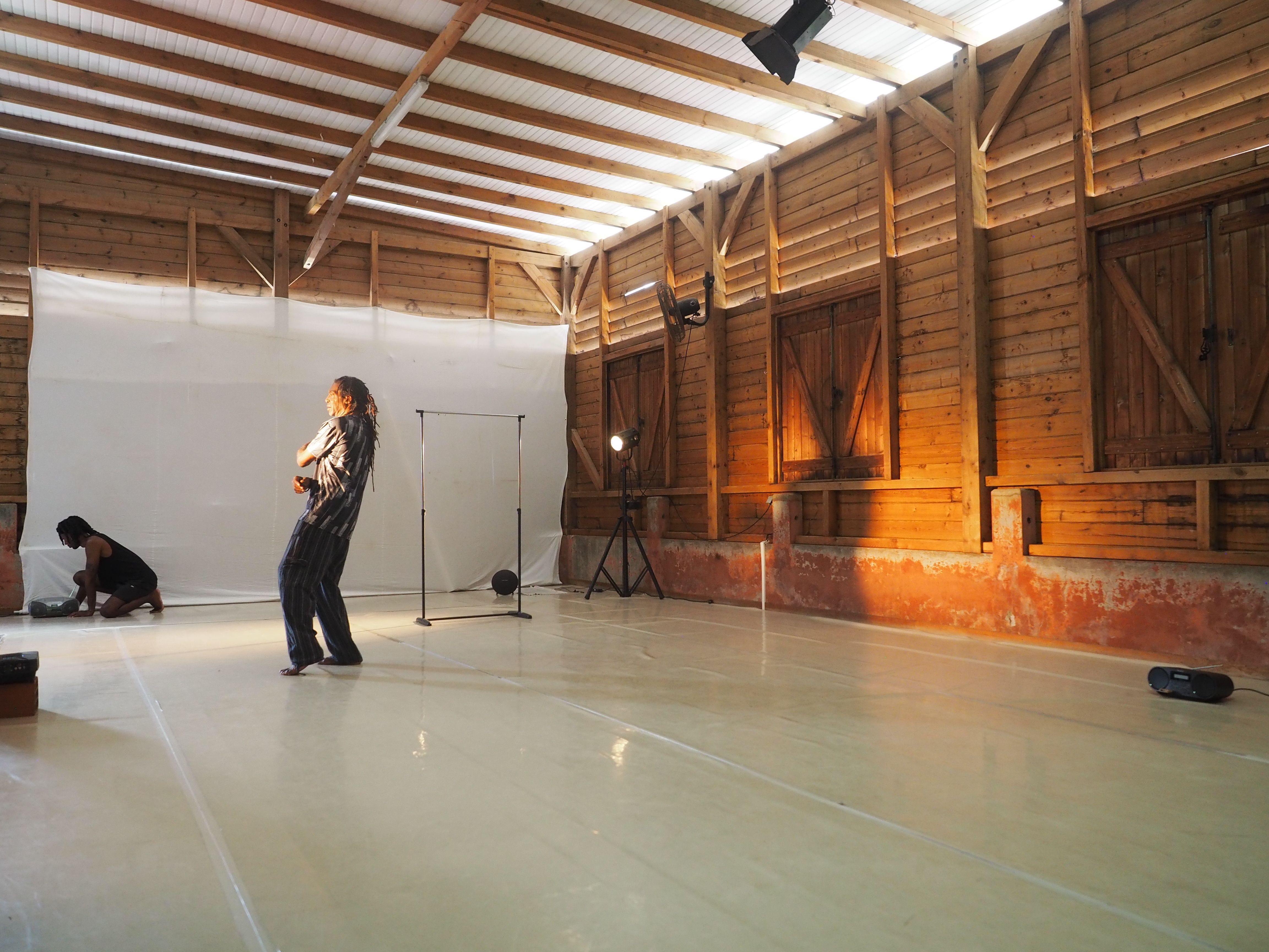 Two people in a rustic wooden room with a white backdrop and studio lights; one stands center, another kneels left.