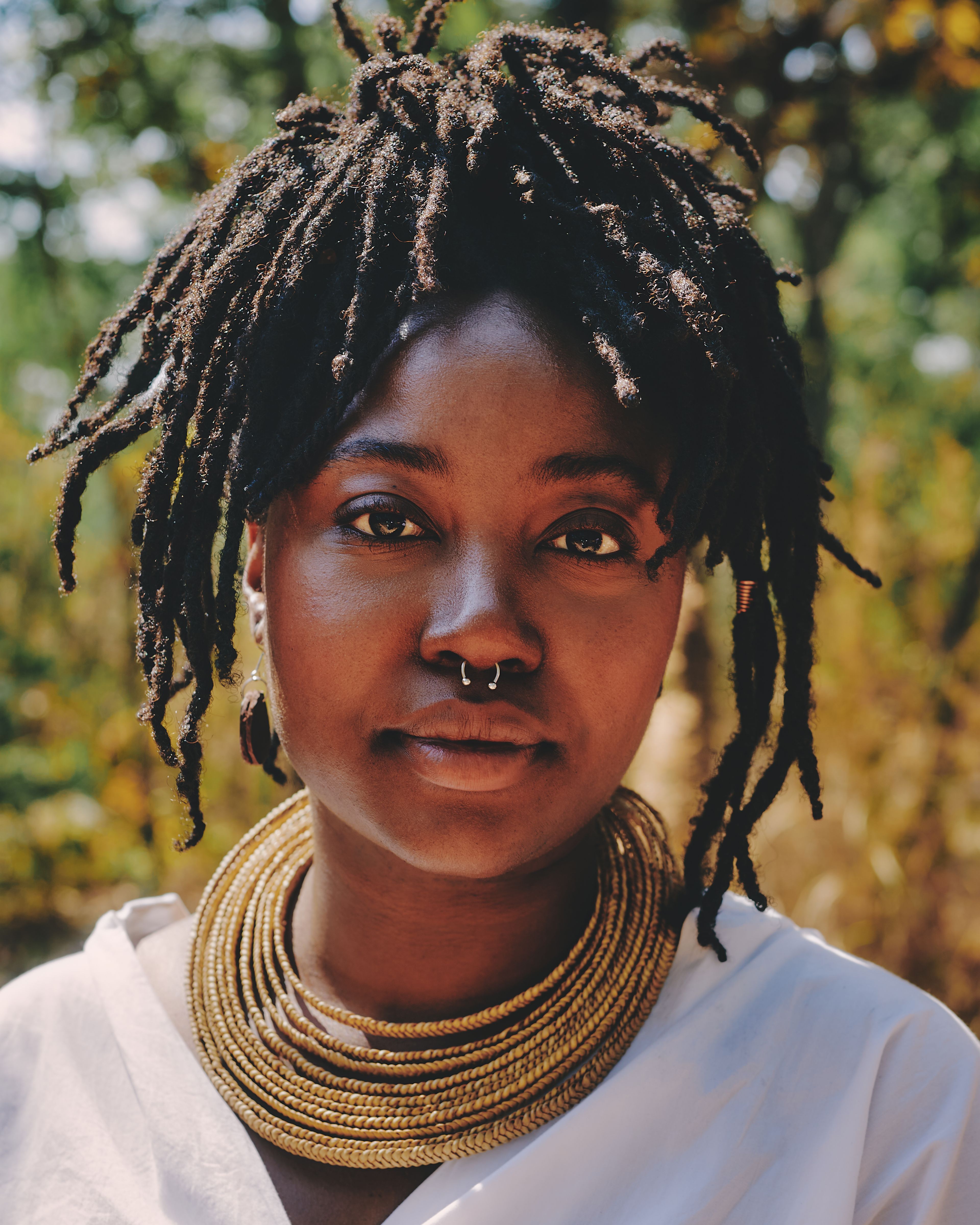 Close-up portrait of a Black person with dreadlocks, a septum ring, and a multi-strand gold necklace, wearing a white shirt outdoors.