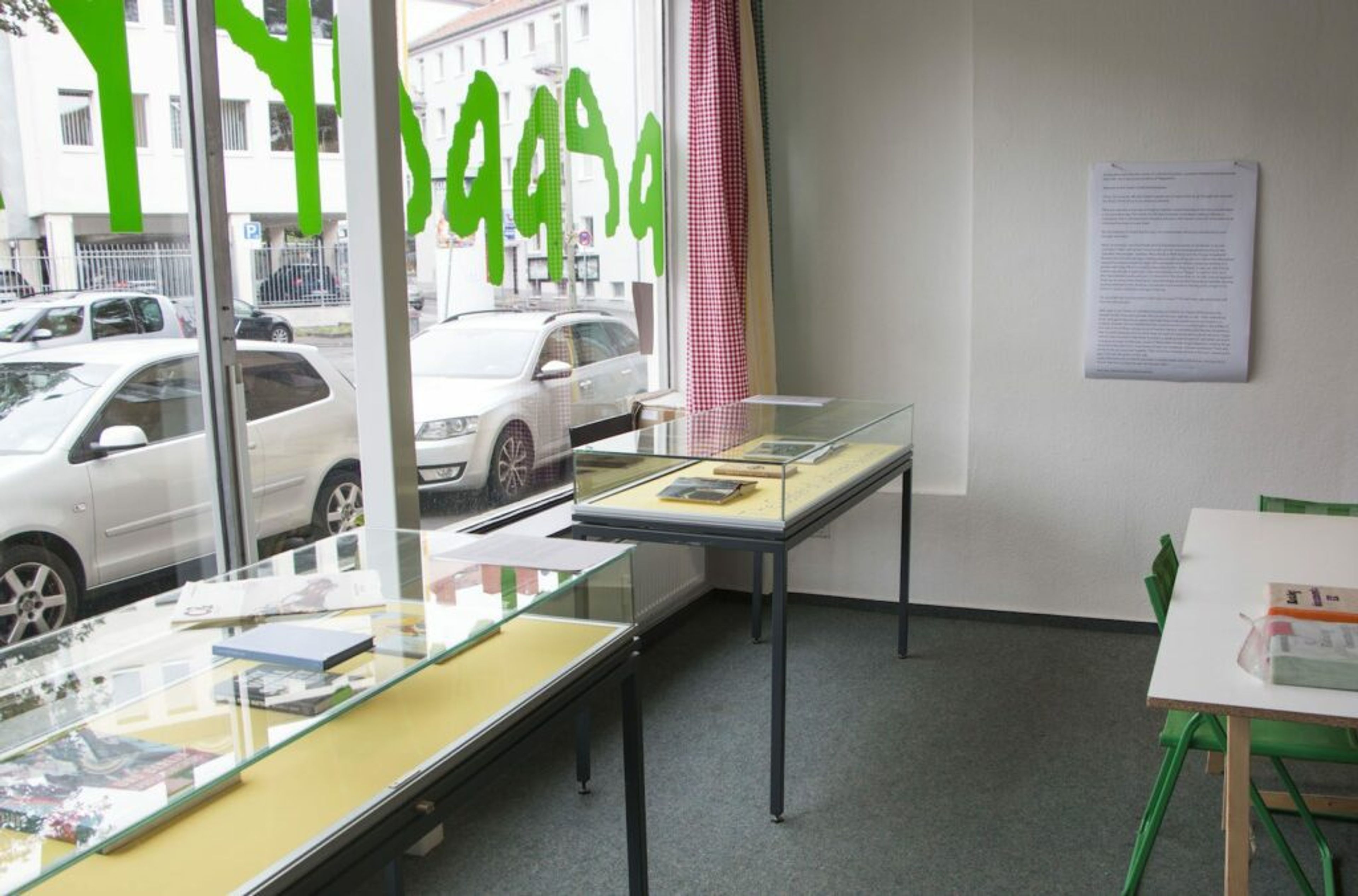 Interior of a room featuring display cases and a table with a green chair. A large window with green text on the glass offers a view of a street with parked cars.