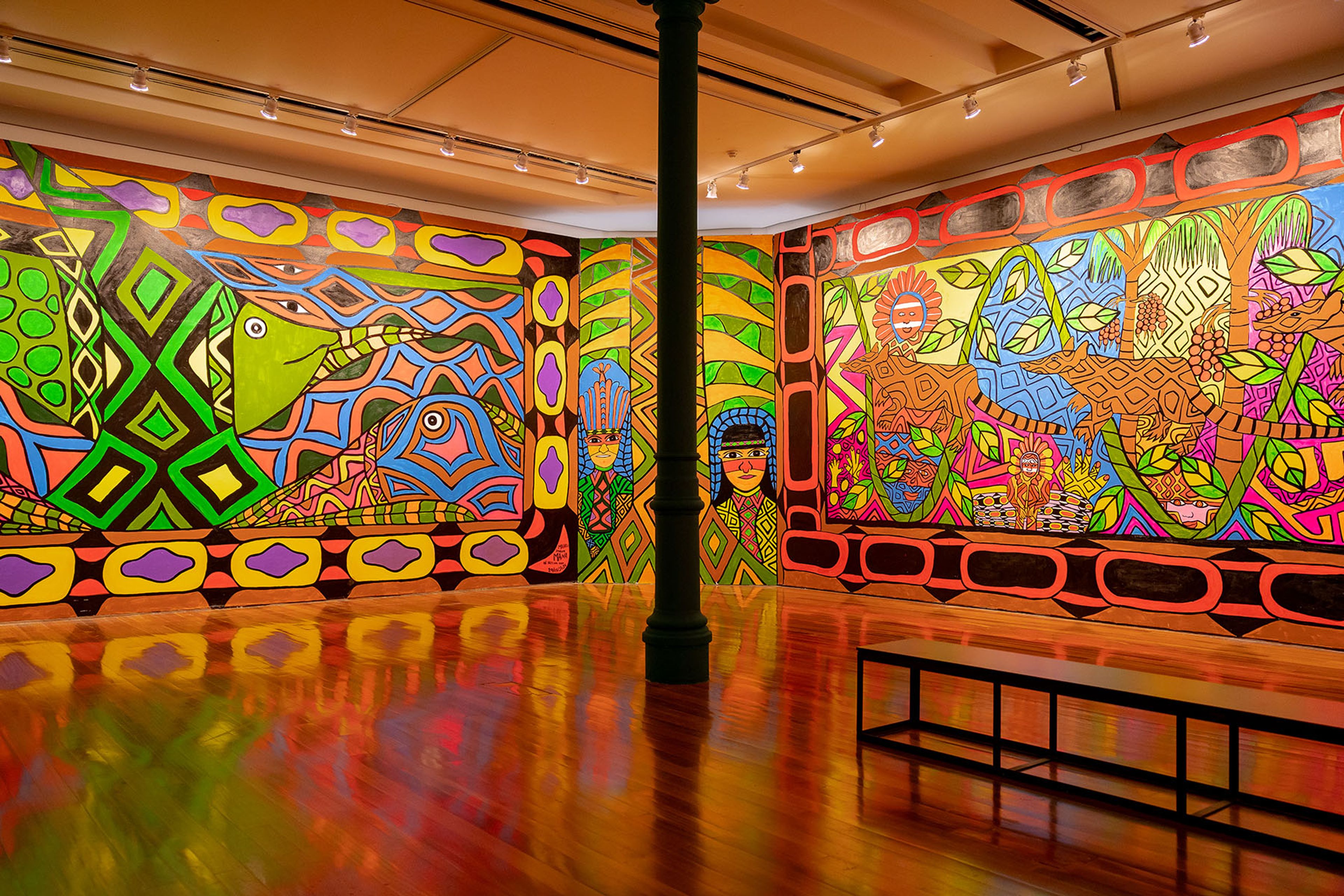Vibrant, intricate murals depicting geometric patterns, stylized animals, and human figures cover art gallery walls, reflected on a glossy wooden floor.