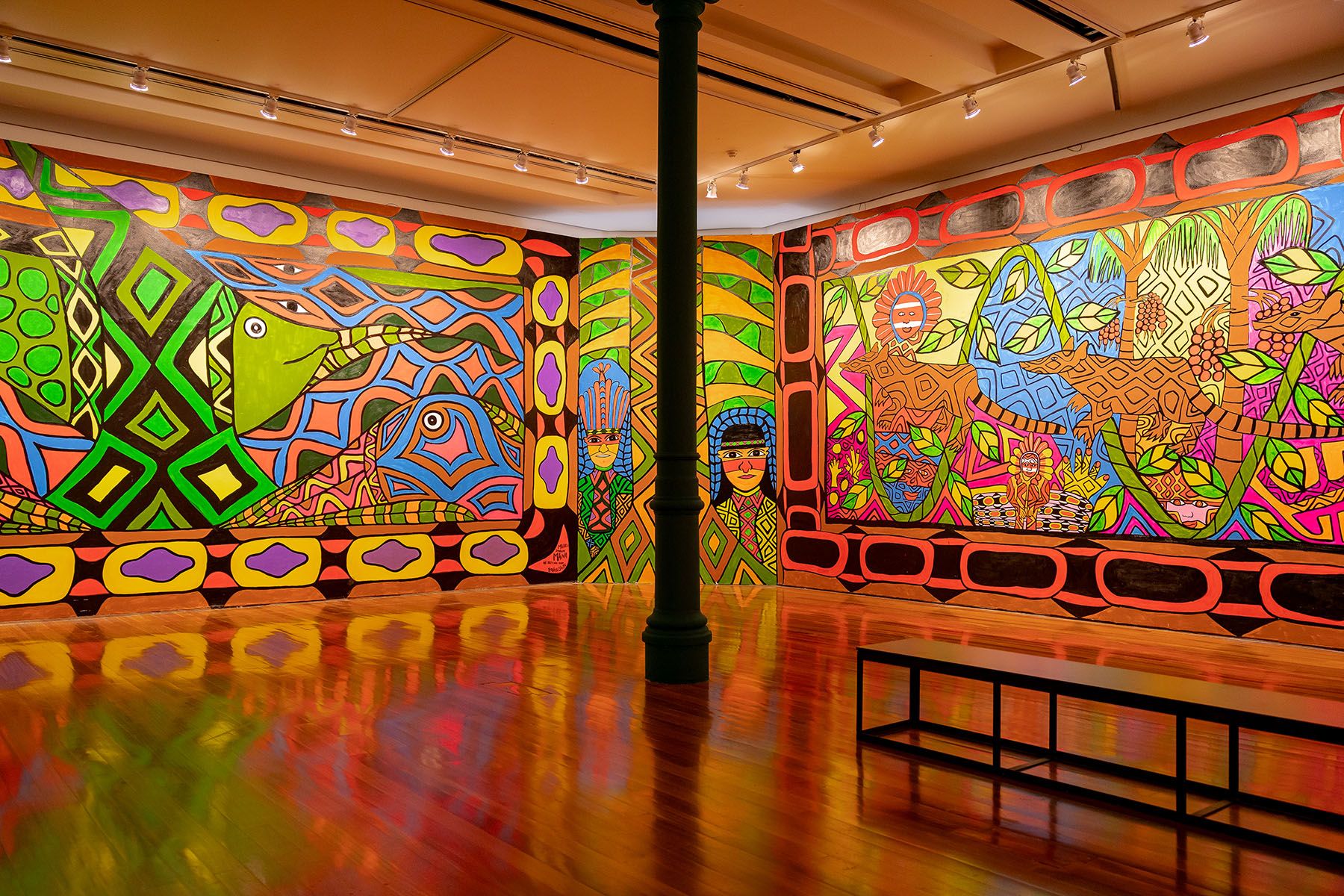 Vibrant, intricate murals depicting geometric patterns, stylized animals, and human figures cover art gallery walls, reflected on a glossy wooden floor.