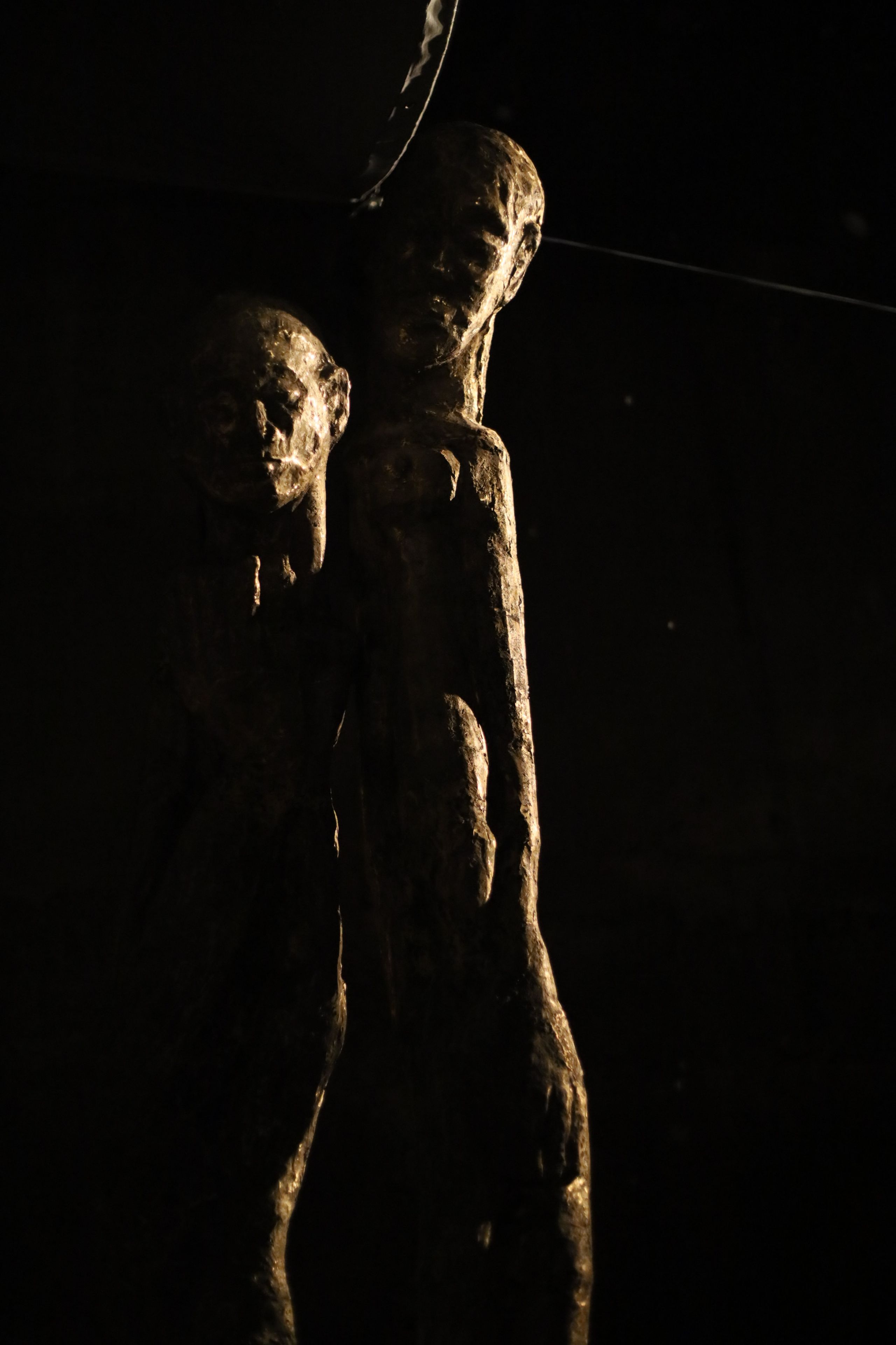 Two textured sculptures of human figures are dimly lit against a dark background.