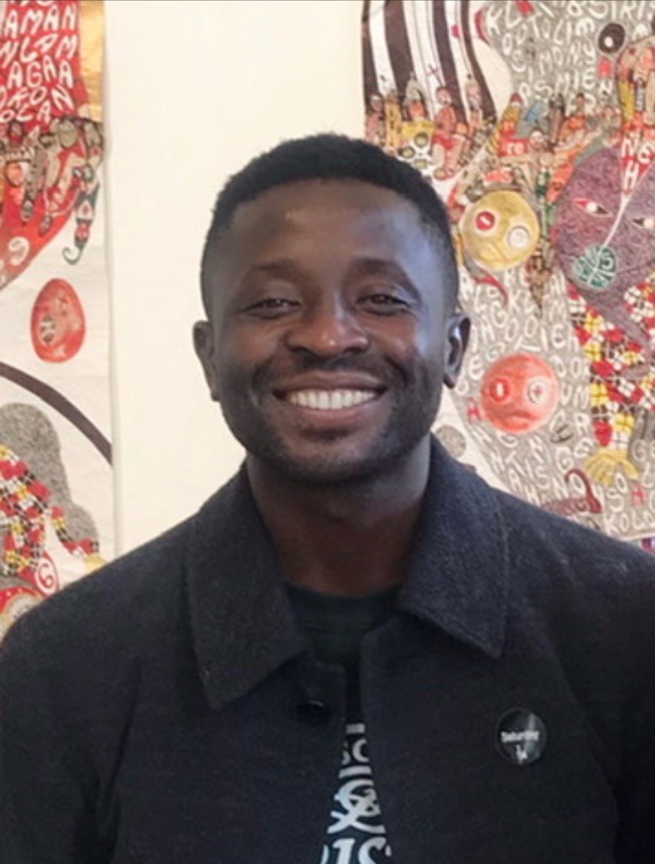 A smiling Black man in a dark coat stands in front of colorful, patterned artworks.