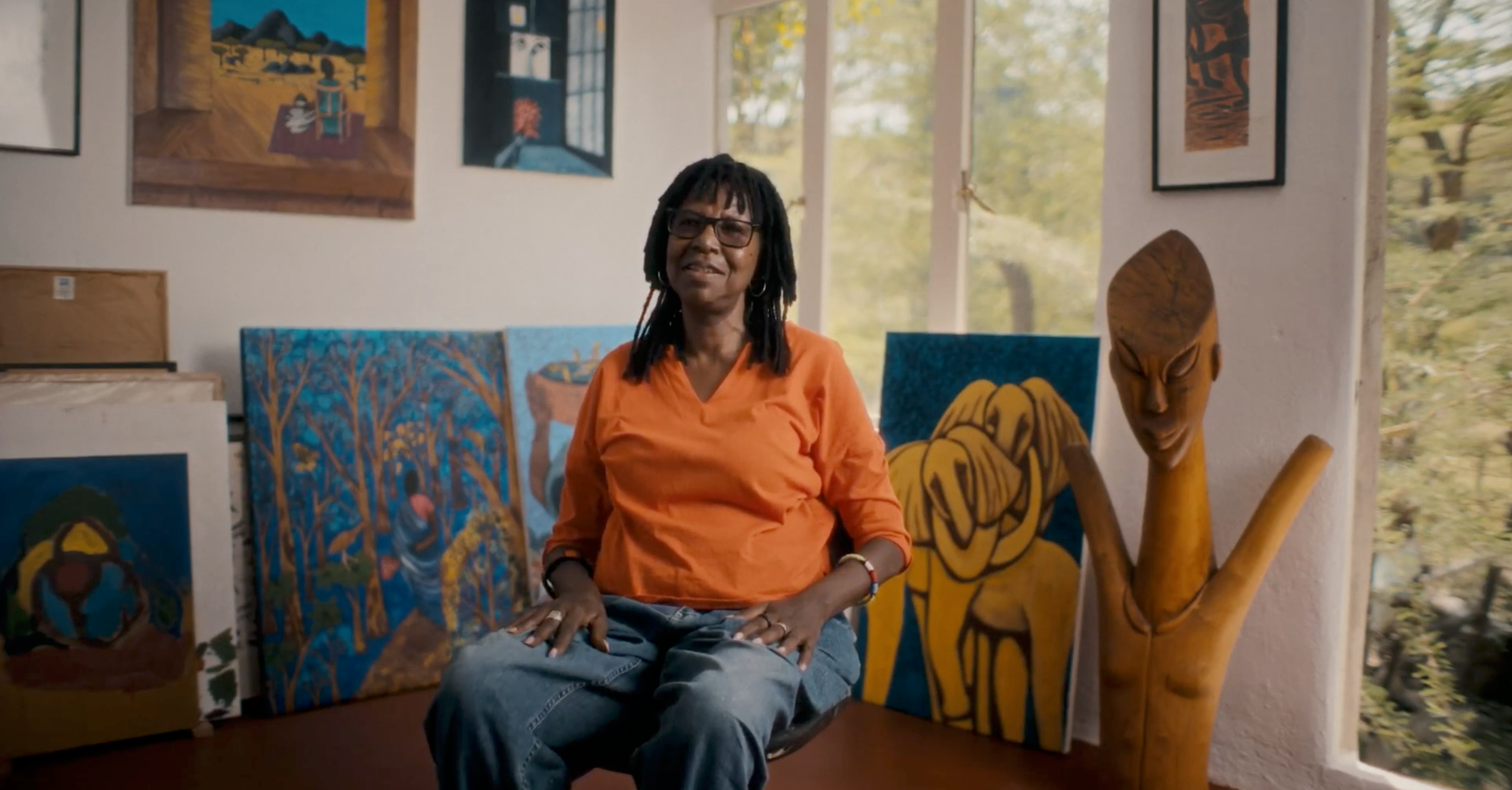 A smiling Black woman with dreadlocks and glasses sits in an art-filled room surrounded by paintings and a wooden sculpture.