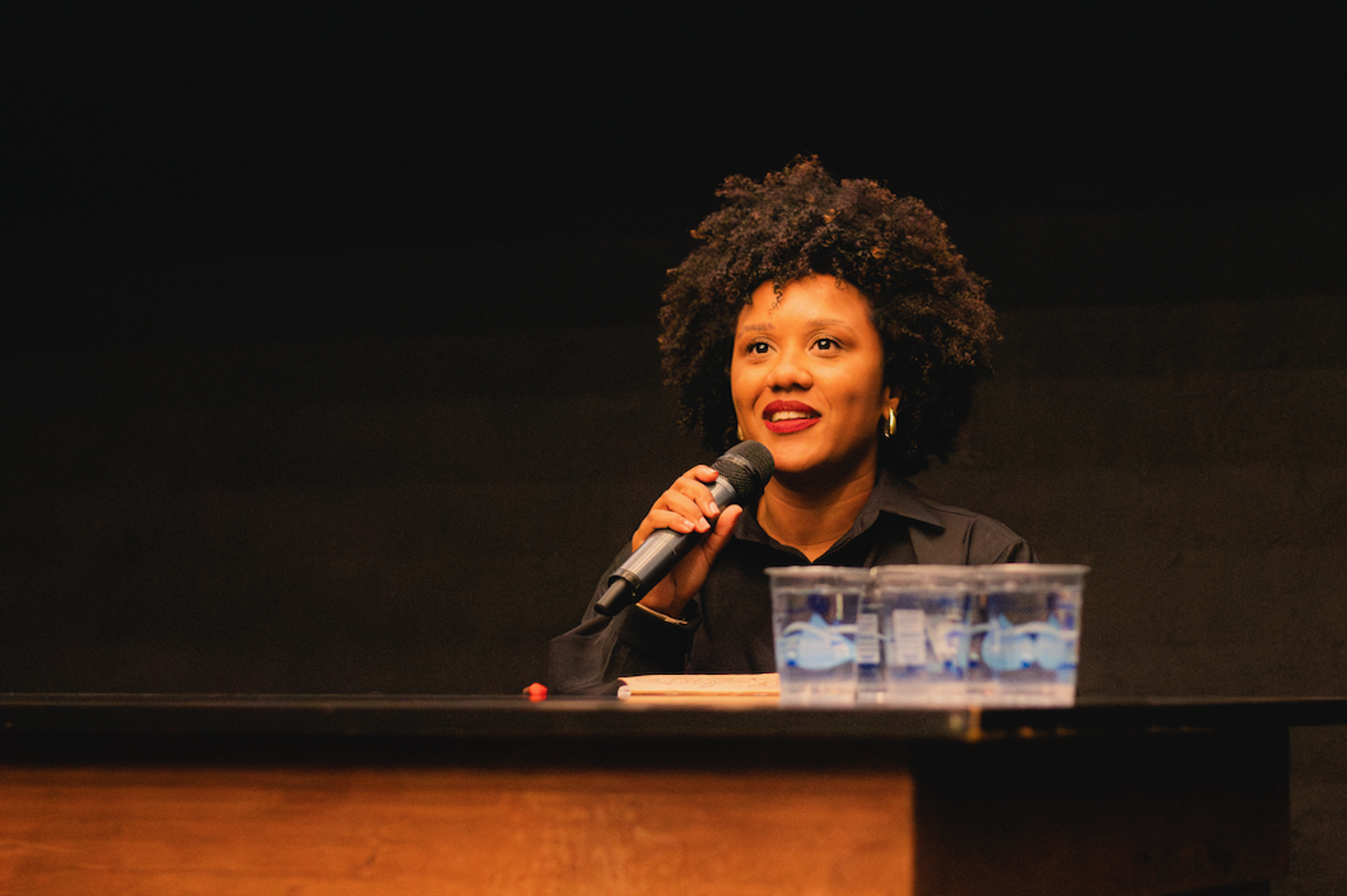 A Black woman with curly hair speaks into a microphone from behind a table.