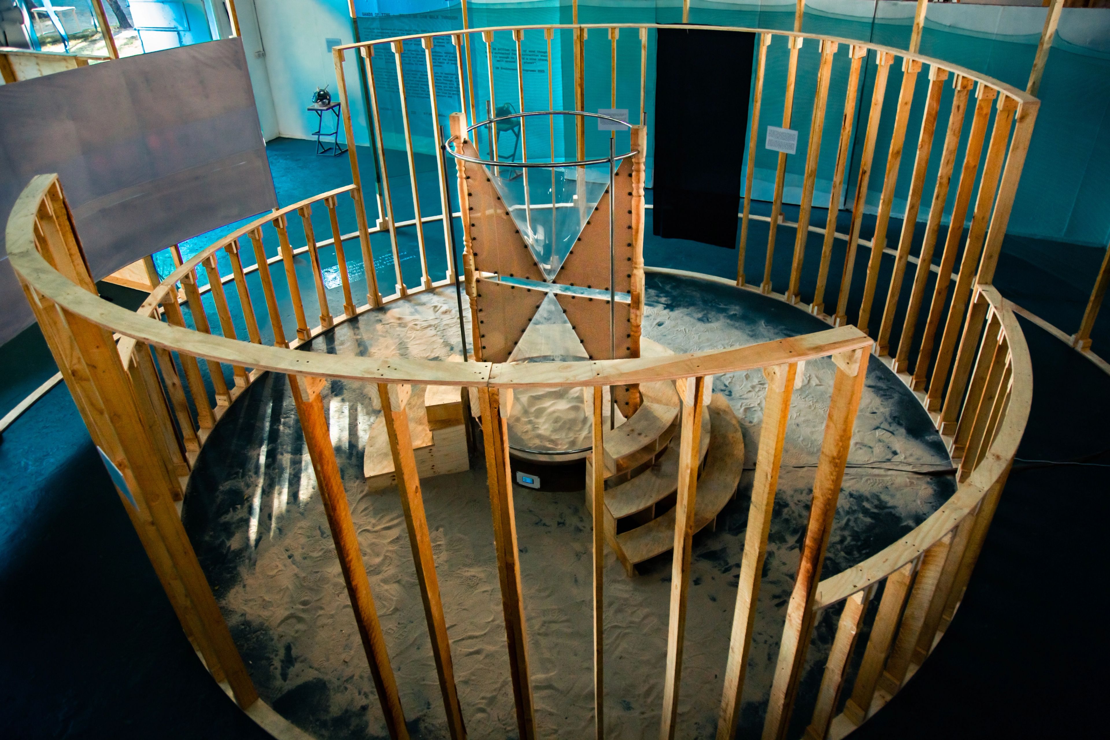 A circular wooden enclosure with sand on the ground surrounds a large central hourglass-shaped sculpture and a small spiral staircase.