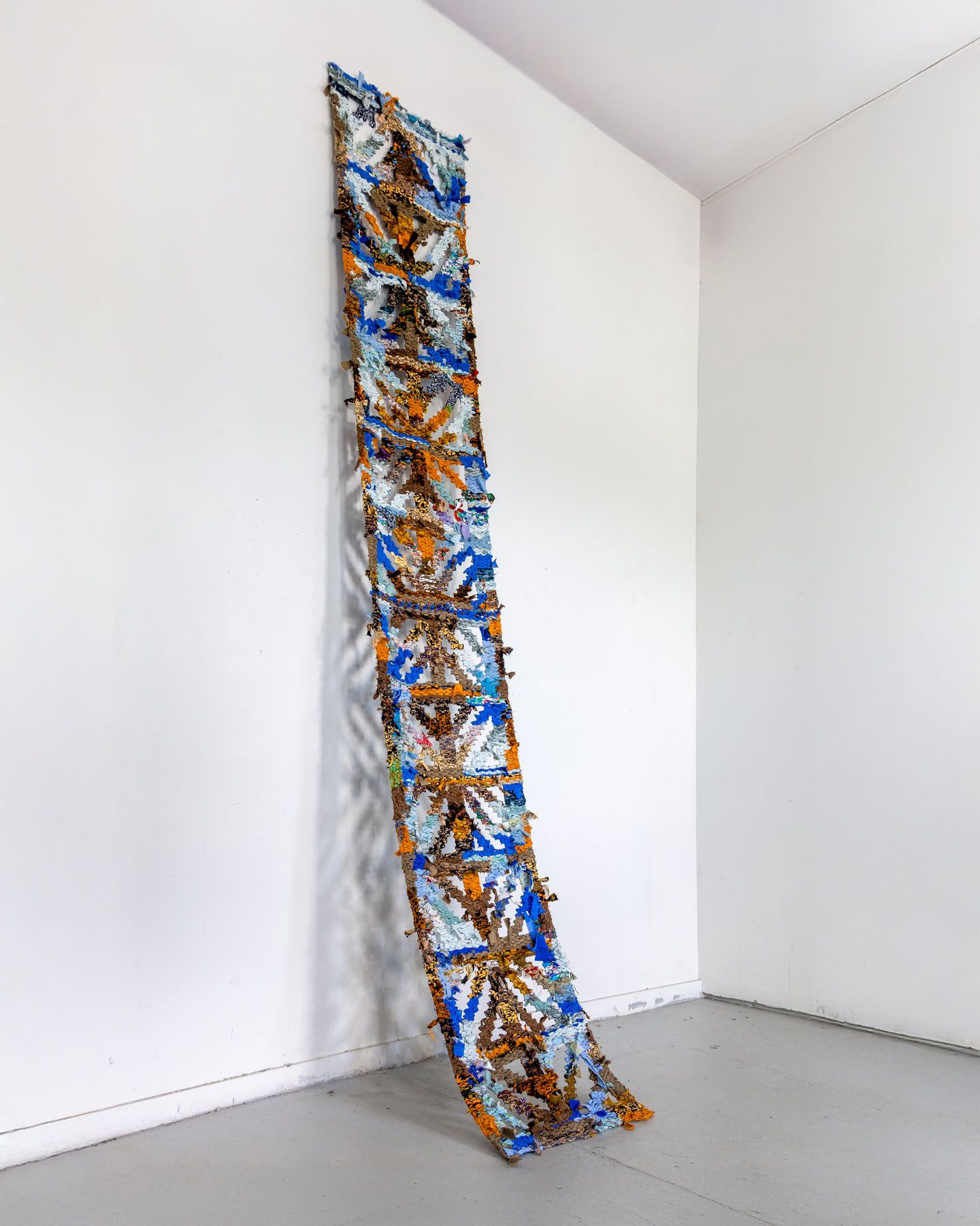 Long, narrow textile artwork made of colorful fabric scraps in blue, orange, and brown, hanging on a white wall.