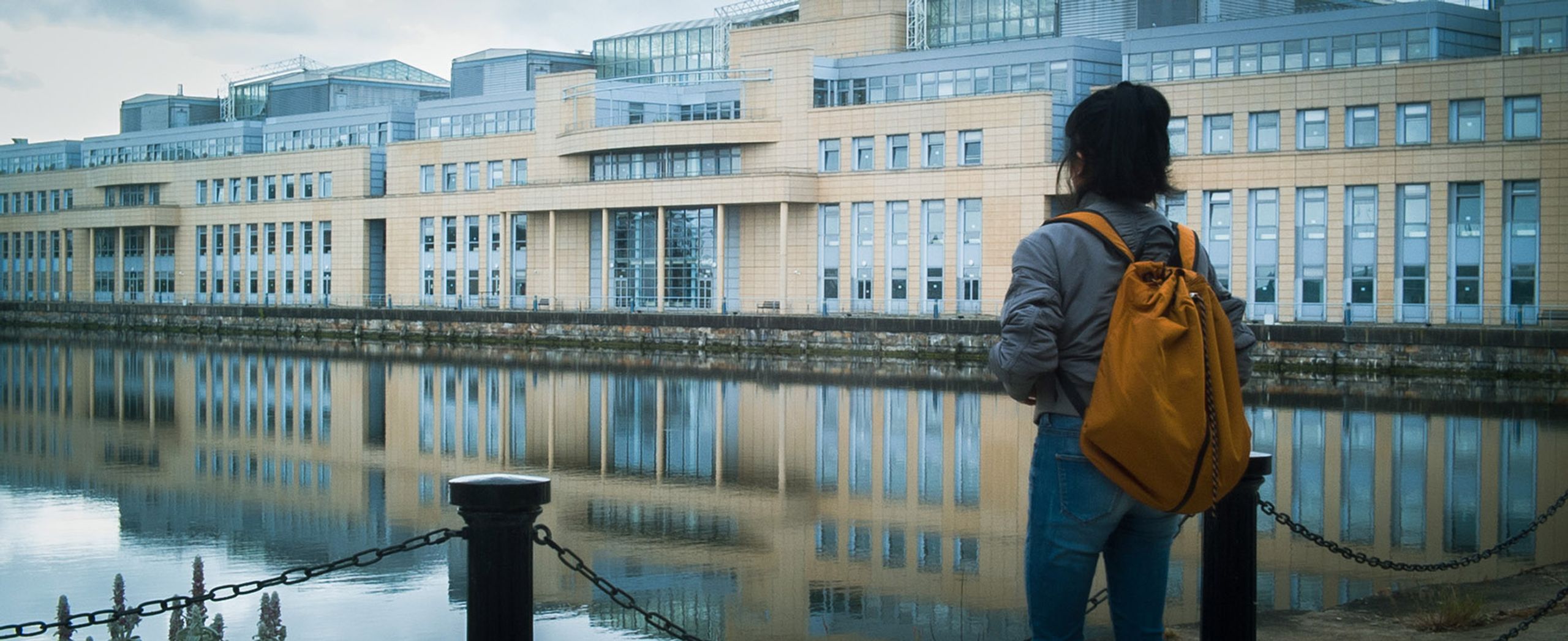 A person with a yellow backpack stands by a waterfront, looking at a modern building reflected in the water.