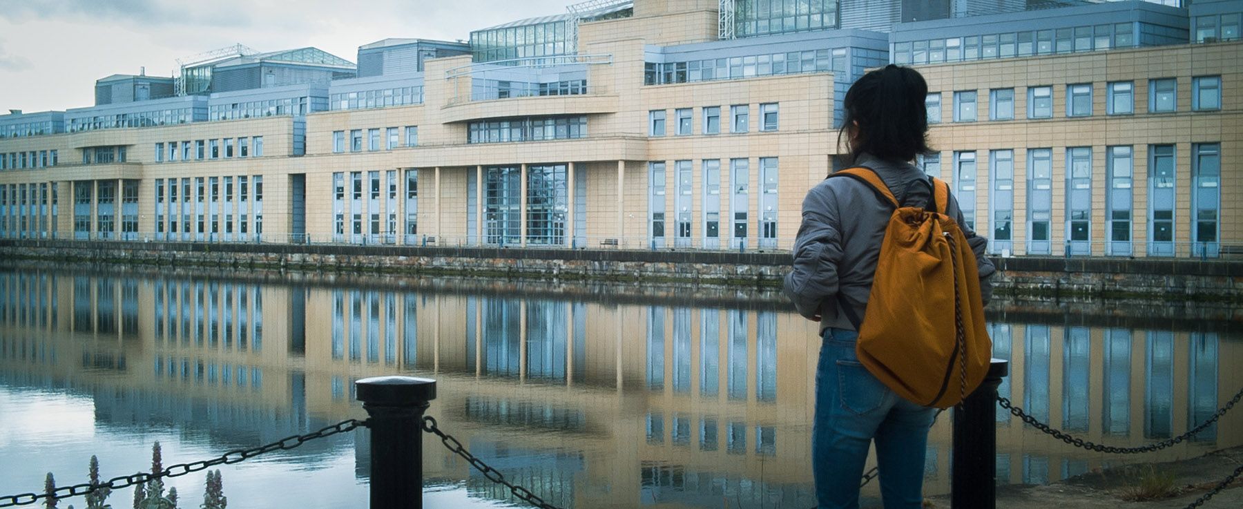 A person with a yellow backpack stands by a waterfront, looking at a modern building reflected in the water.