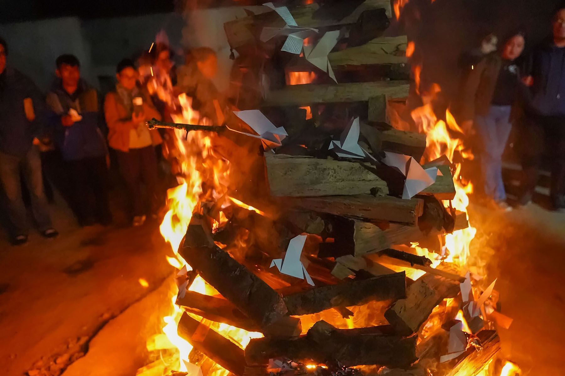 A large bonfire with paper offerings burning, surrounded by people at night.