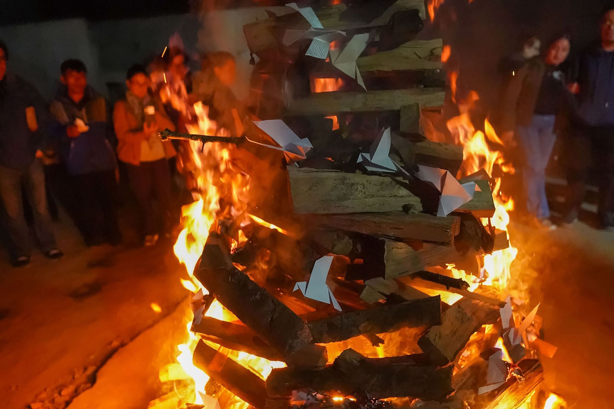 A large bonfire with paper offerings burning, surrounded by people at night.