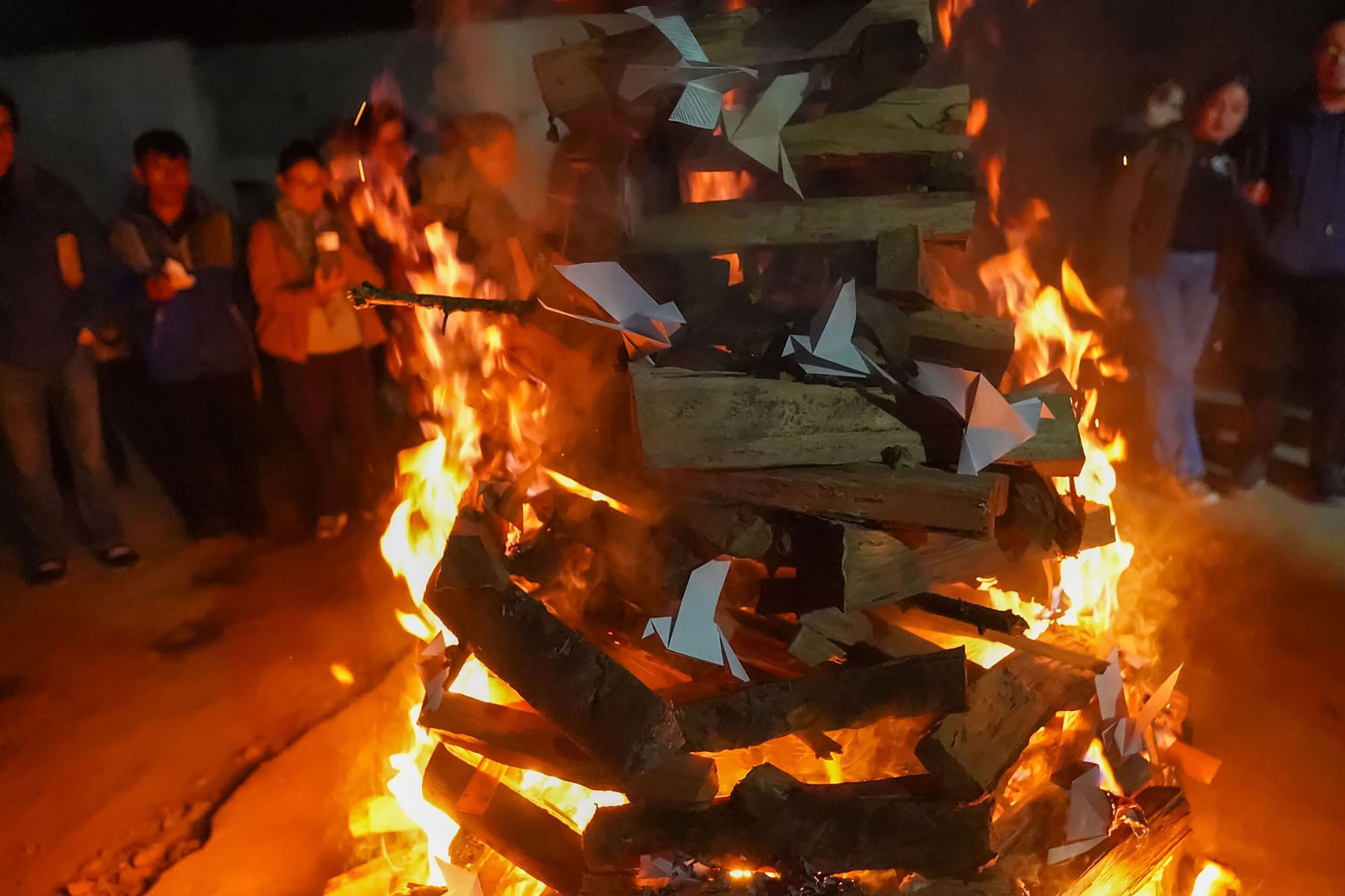 A large bonfire with paper offerings burning, surrounded by people at night.