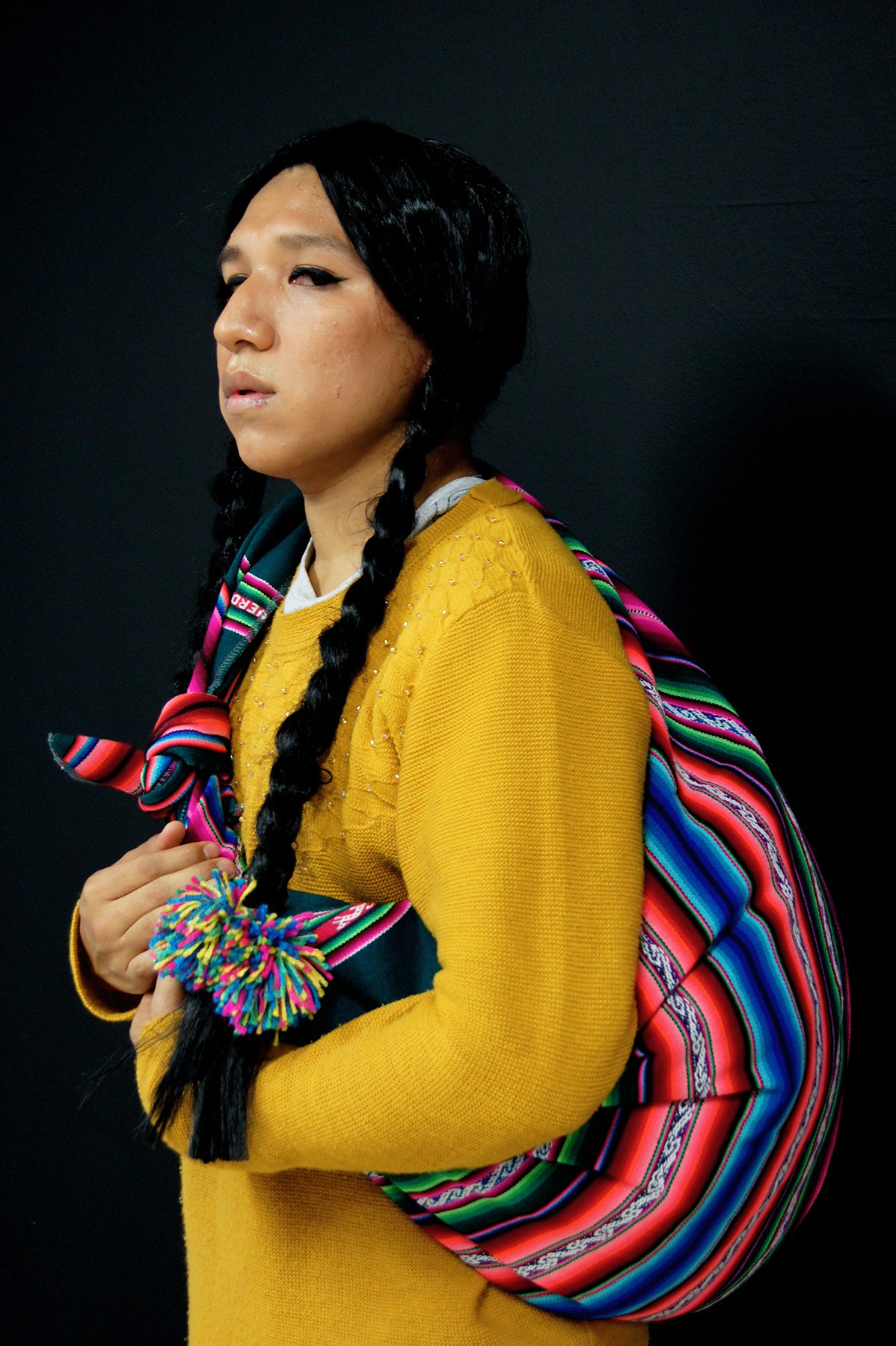 Person with braided pigtails wearing a yellow sweater and a colorful striped bag with a pompom over their shoulder.
