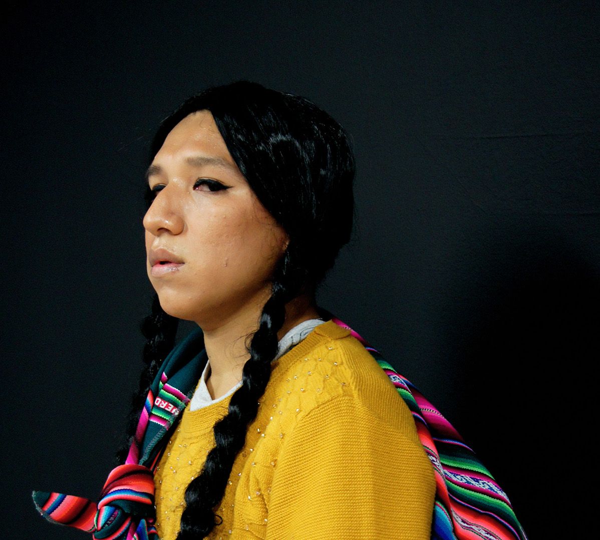 Person with braided pigtails wearing a yellow sweater and a colorful striped bag with a pompom over their shoulder.