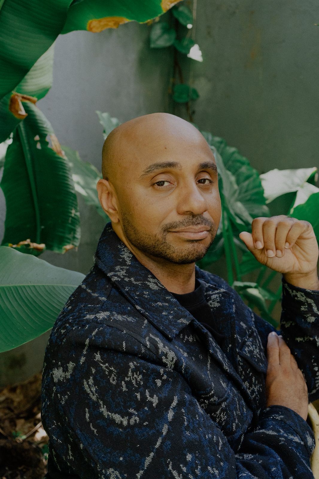 A bald man with a light beard, wearing a dark patterned jacket, looks directly at the camera while surrounded by lush green foliage.
