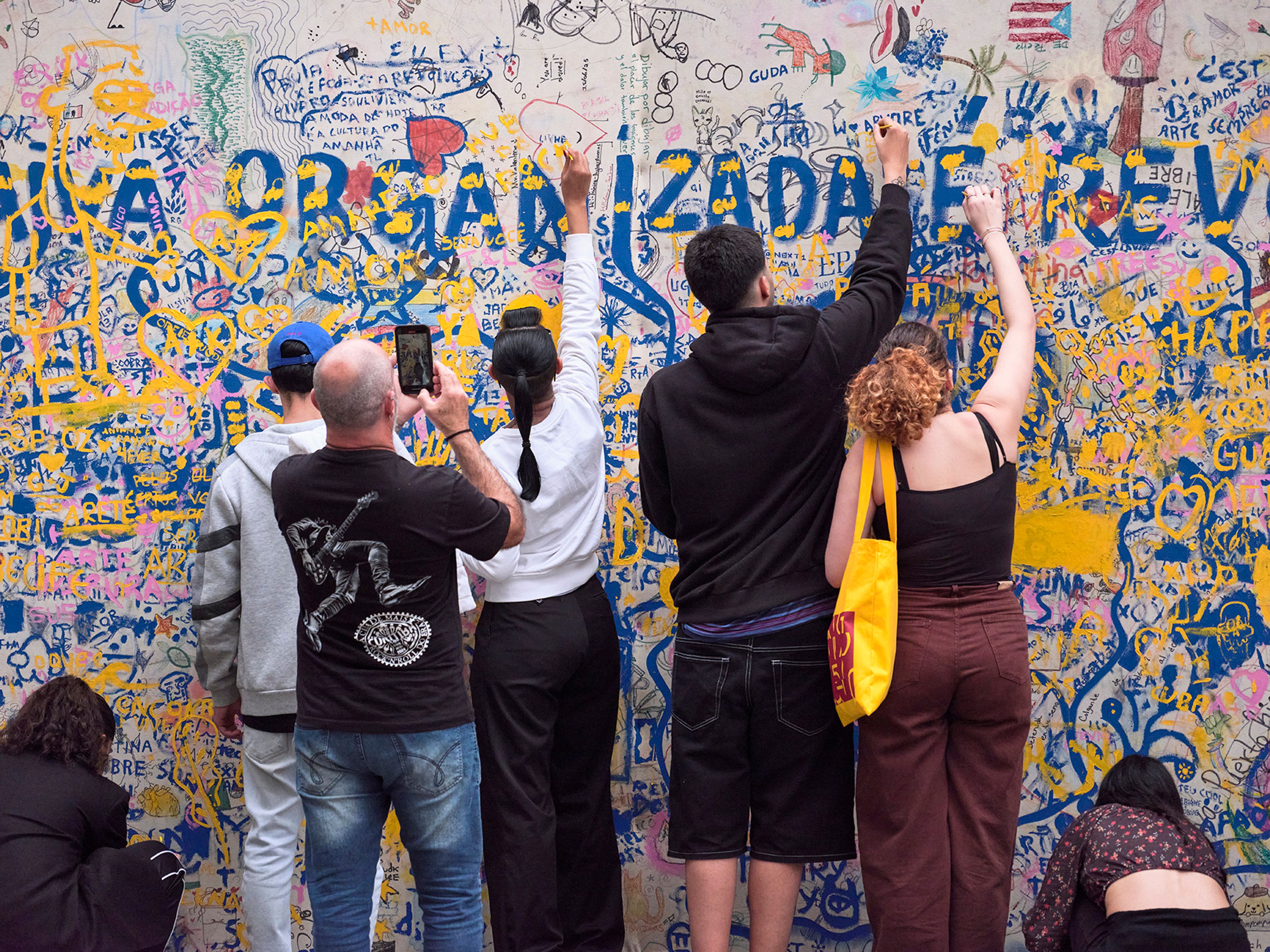 A group of people write on and photograph a wall covered in colorful, layered graffiti.