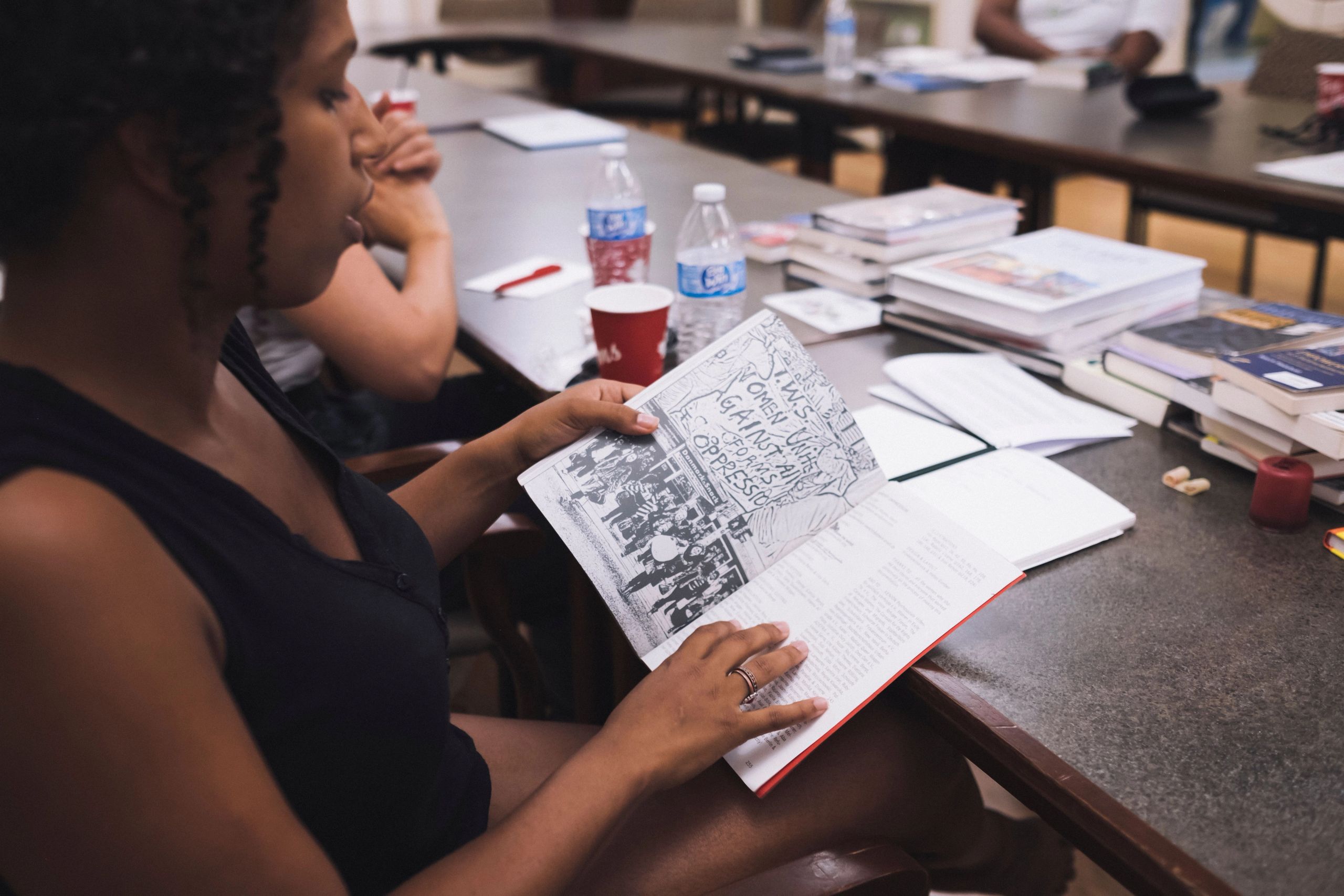 A woman with curly hair reads an open book featuring a protest image and text, sitting at a table with other books.
