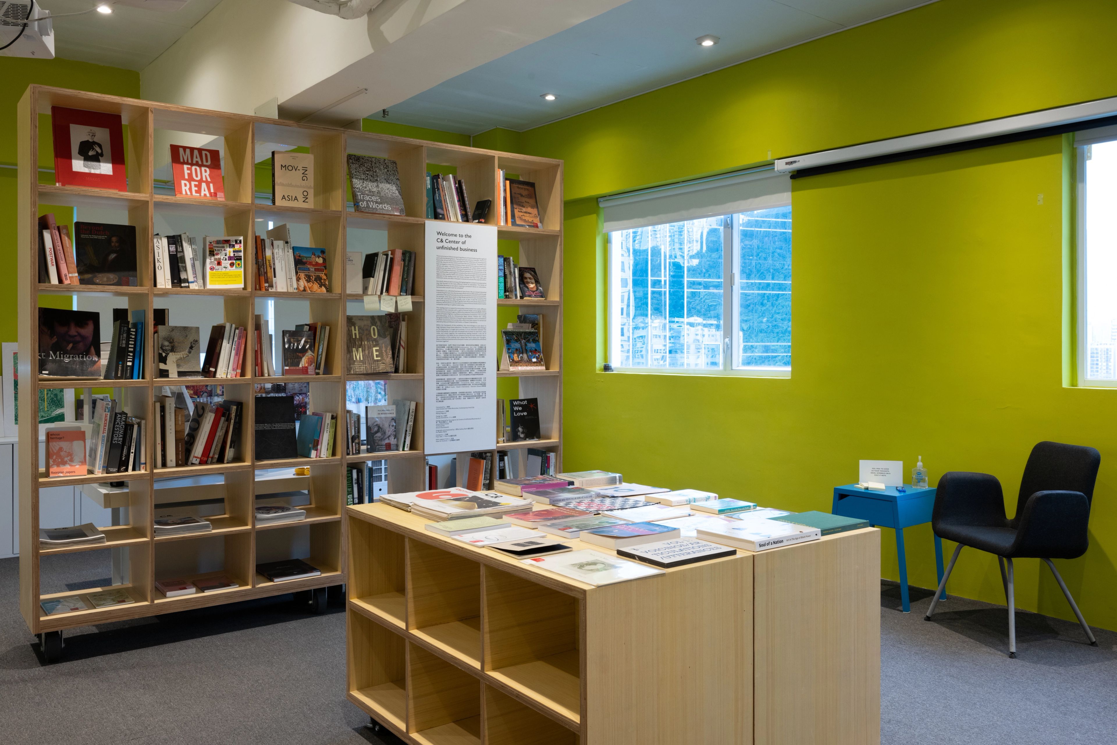 A lime green room with a wooden bookshelf full of books, a central display table with books, a window, and a chair.