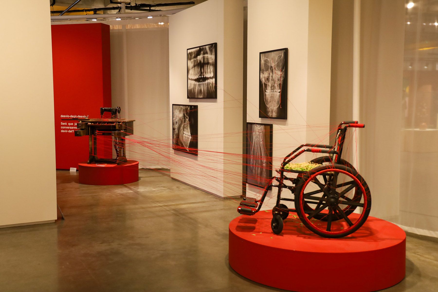 Art installation: a black wheelchair with red accents and a sewing machine, both on red pedestals, connected by red threads. X-ray images are on the wall.