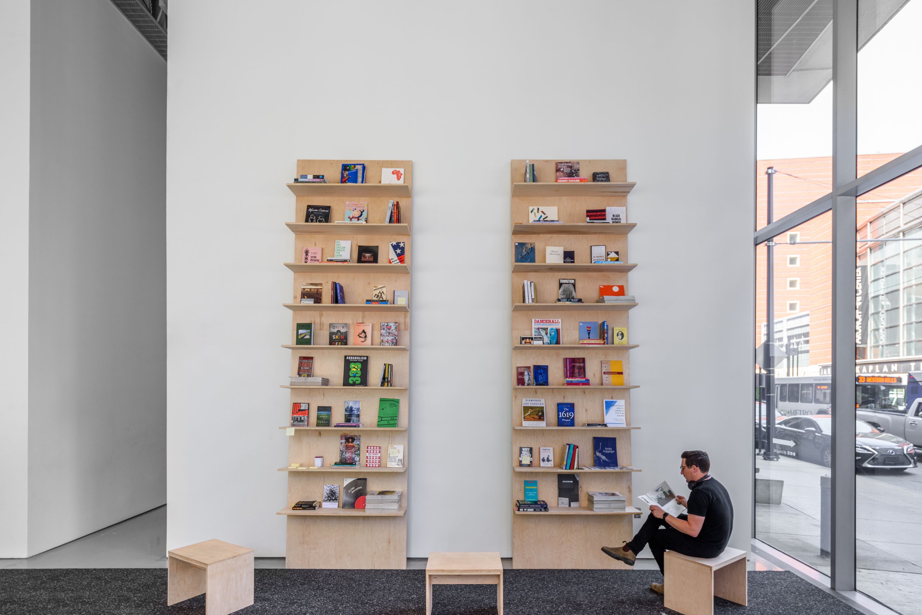 Man reading on a stool by two minimalist light wood bookshelves in a modern room with a large window view of a city street.