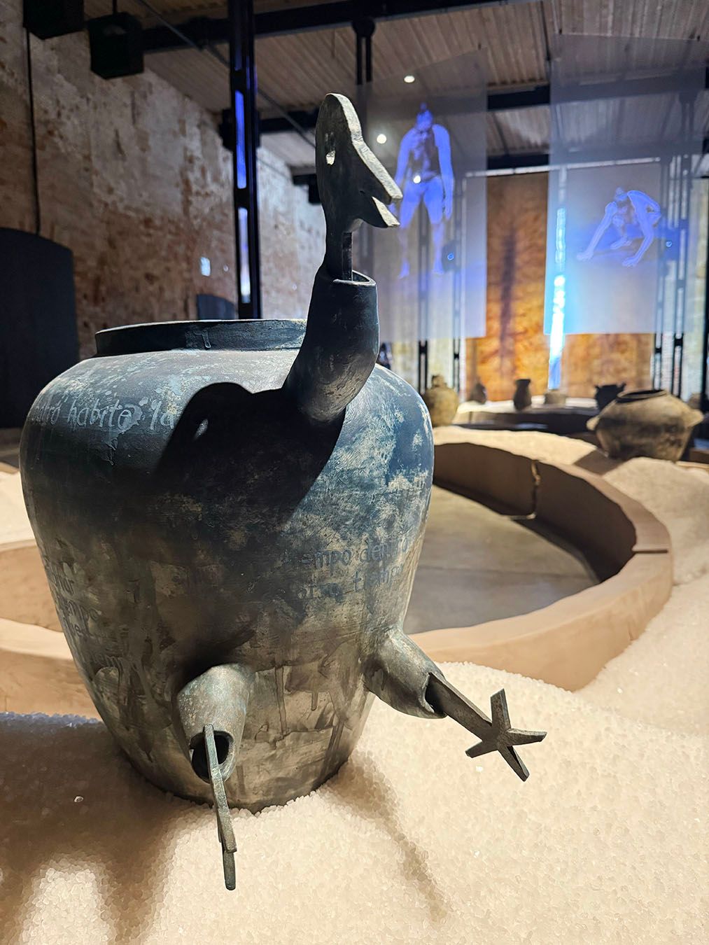 A dark, sculptural metal pot with a bird-shaped handle and a star-shaped adornment sits in white granular material in an exhibition space with projected blue figures in the background.