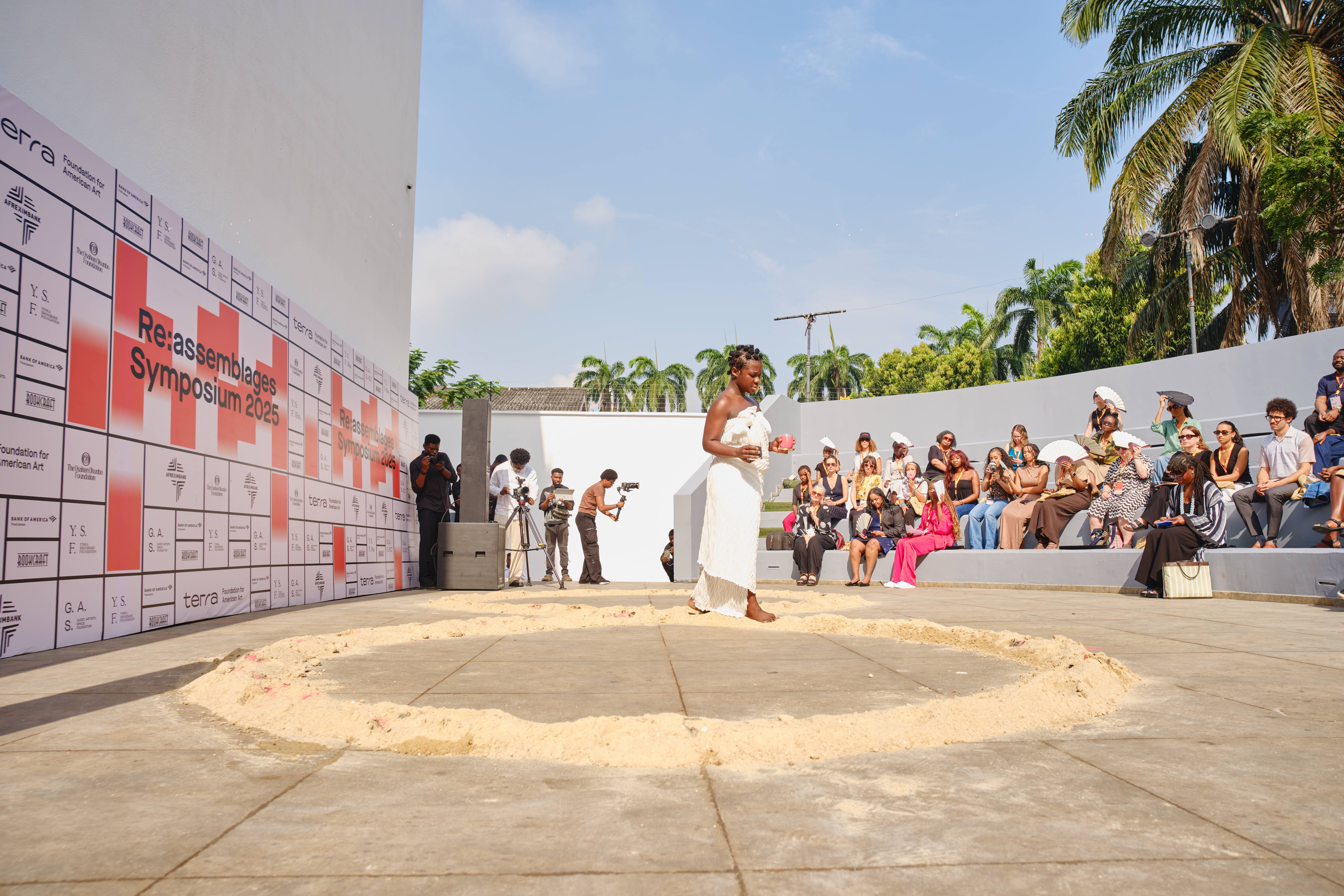 A Black woman in a white dress stands in a sand circle, speaking to an audience seated on tiered steps at an outdoor "Re:assemblages Symposium 2025" event.