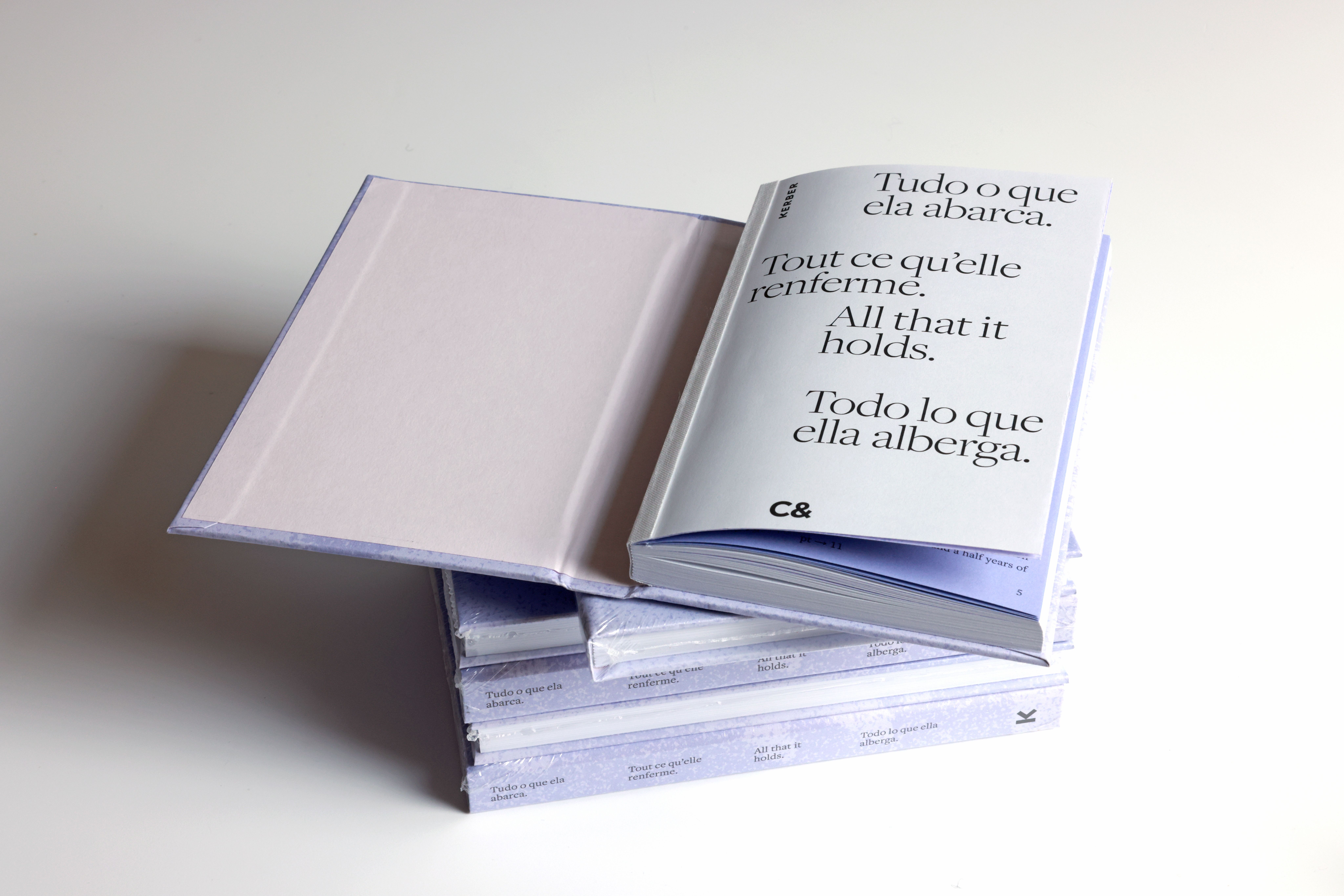 A stack of light purple books, with one open displaying "All that it holds" in multiple languages.