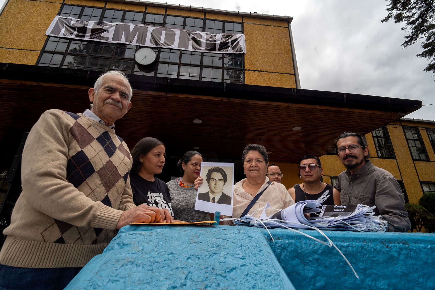 A group of people stand outside a building with a "MEMORIA" banner, one person holding a black and white photo of a young man.