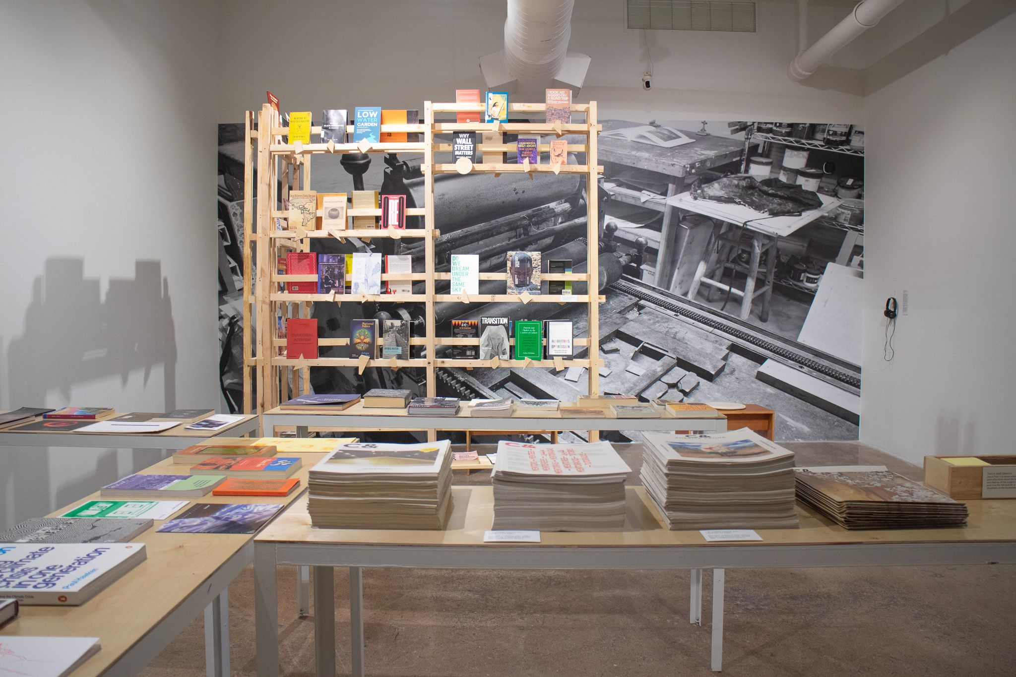 Gallery space with tables displaying publications, a wooden bookshelf with books, and a large black and white mural of a workshop.