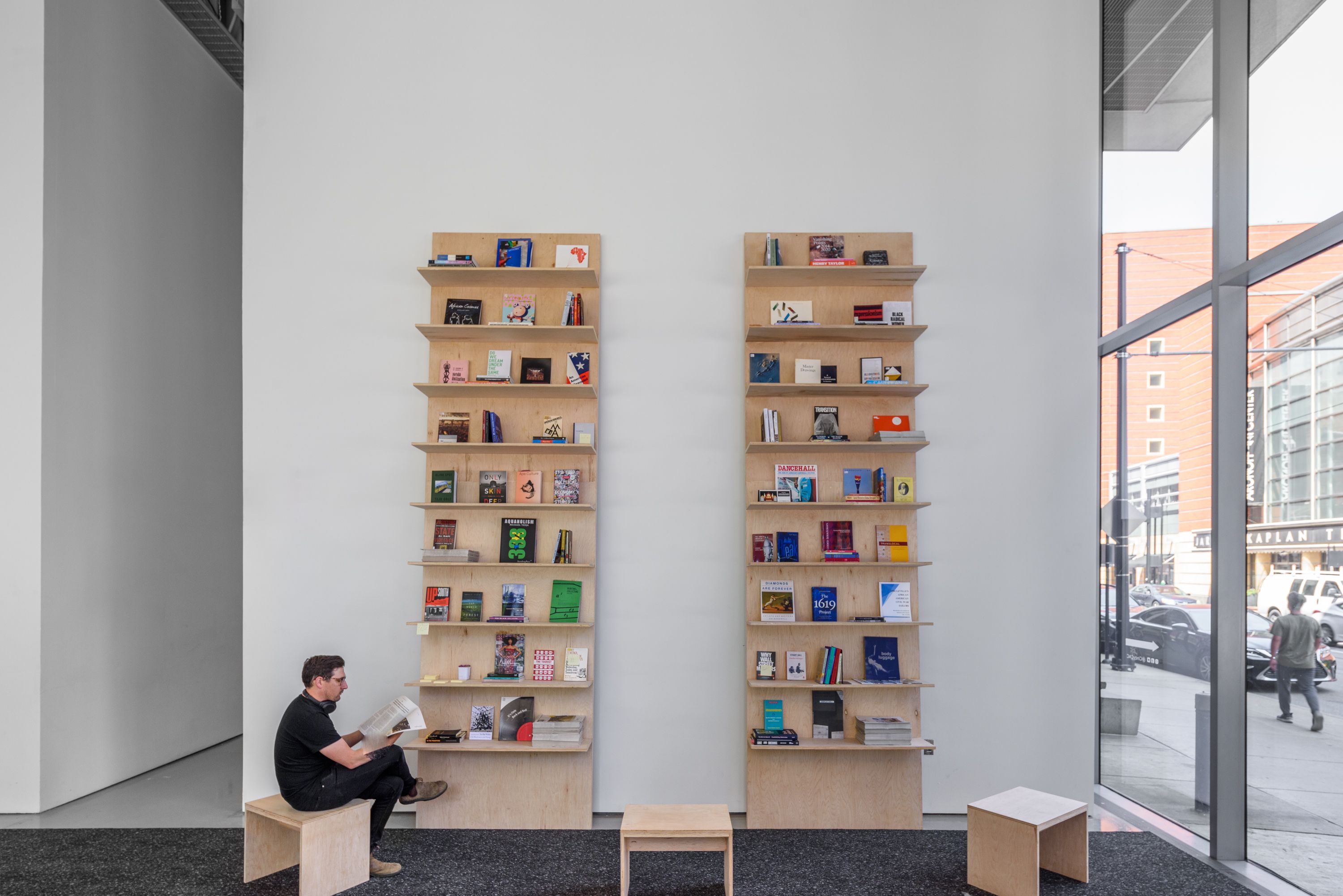 A person reads on a wooden bench between two tall bookshelves in a modern room with large windows.