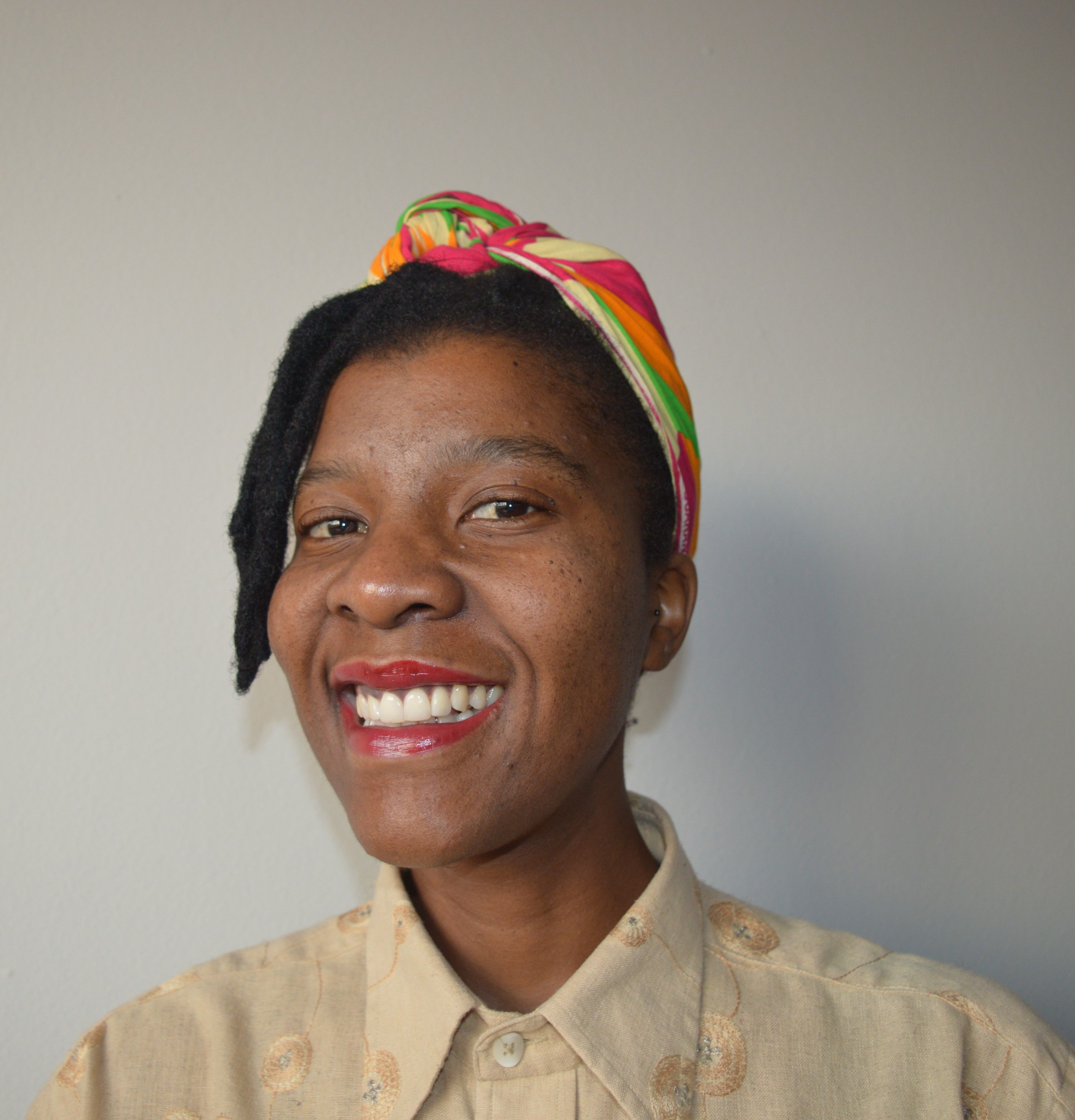 A smiling Black person in a colorful head wrap, wearing red lipstick and a beige patterned shirt.