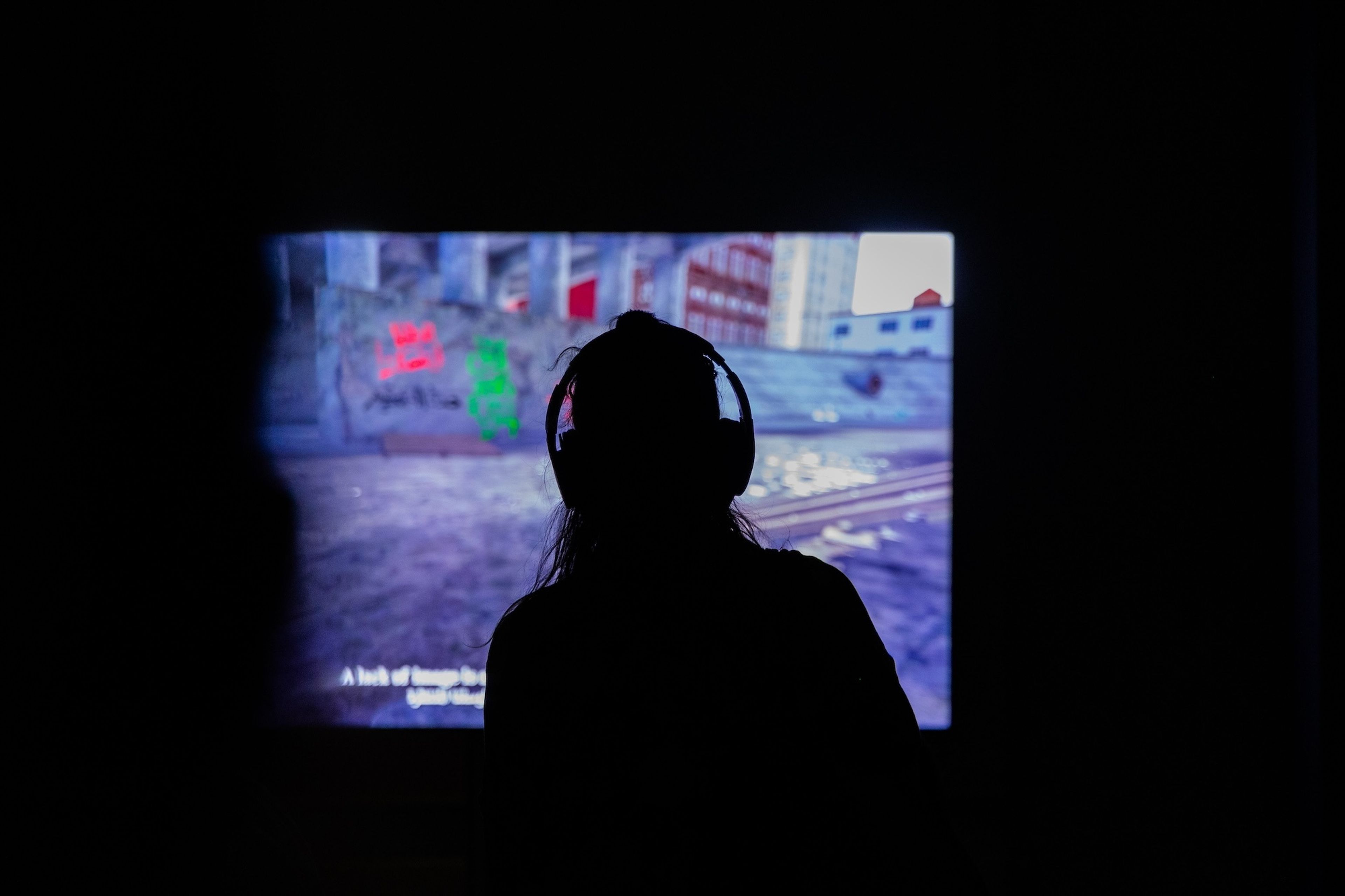 Silhouetted person wearing headphones watches a projected urban scene in a dark room.