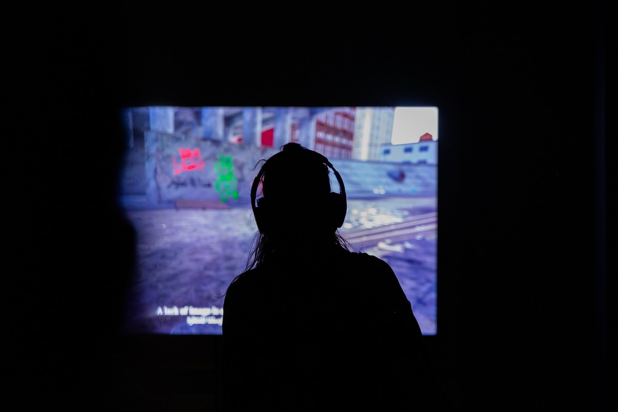 Silhouetted person wearing headphones watches a projected urban scene in a dark room.