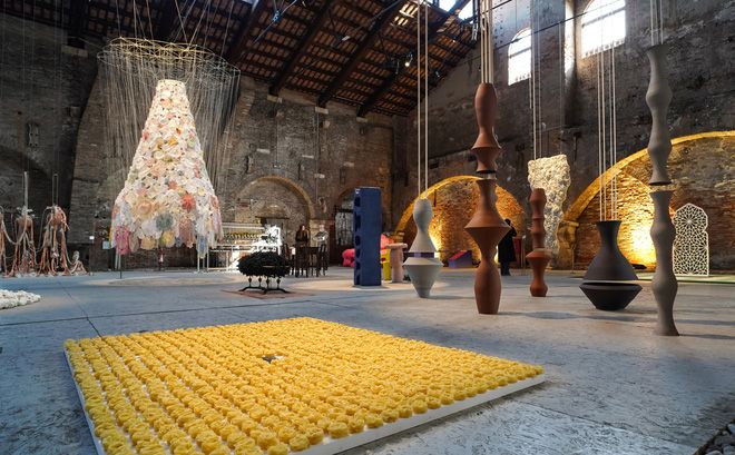 Art exhibition in a large, industrial hall featuring a yellow floor installation, a conical hanging sculpture, and suspended abstract vase-like forms.